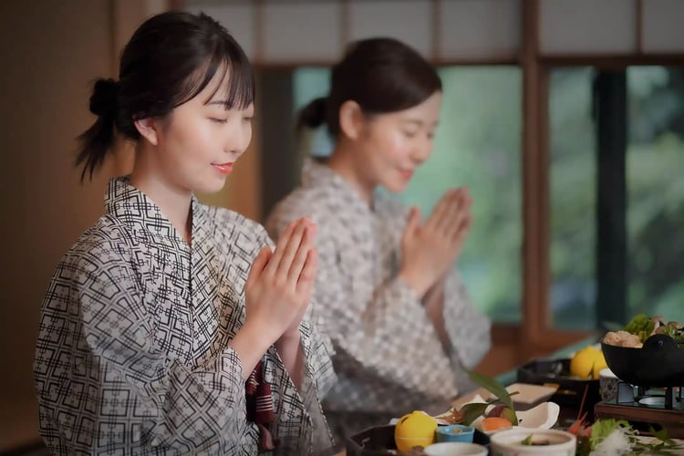 Two women in yukata, dining at a ryokan, close their eyes and put their hands together before eating, saying itadakimasu