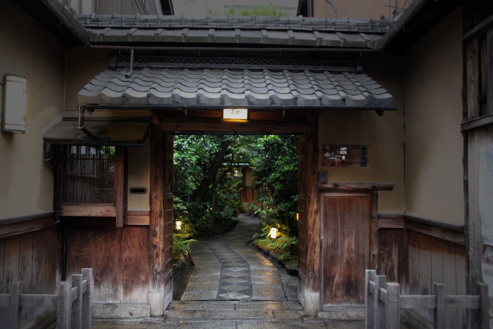 The open wooden gate of a Kyoto ryotei at dusk, a lantern lighting the stone path into a green garden