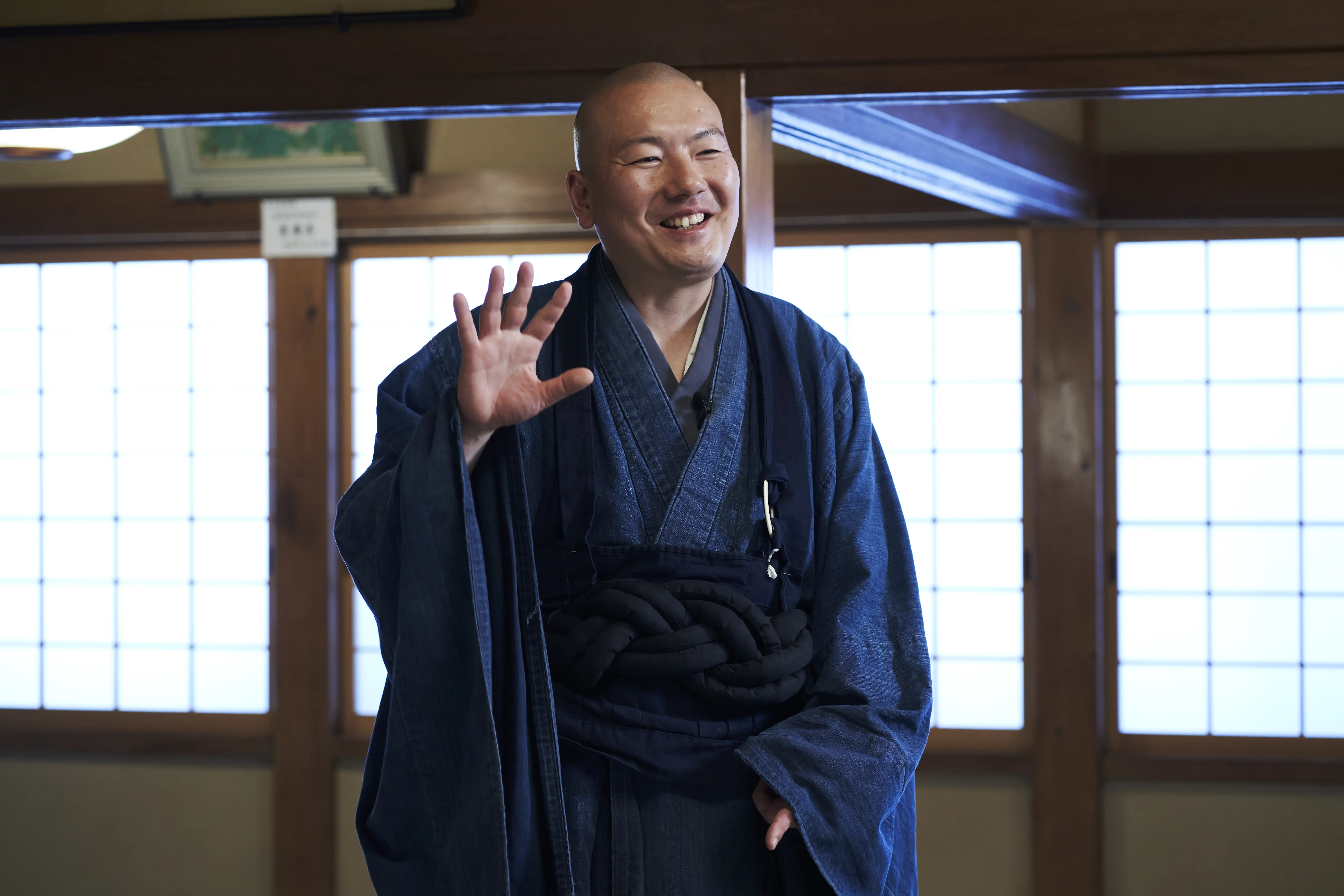 Rev. Taijun Namiki smiling and raising a hand in greeting in a bright room of Kinryuji lined with shoji windows