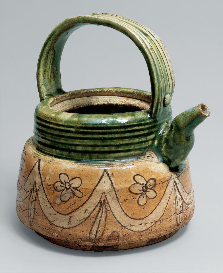 An early seventeenth-century Japanese ewer in the Metropolitan Museum of Art: a ribbed handle arching over the mouth, a deep green glaze across the shoulder, and a buff lower body drawn in dark brown line with a swagged curtain and five-petalled blossoms