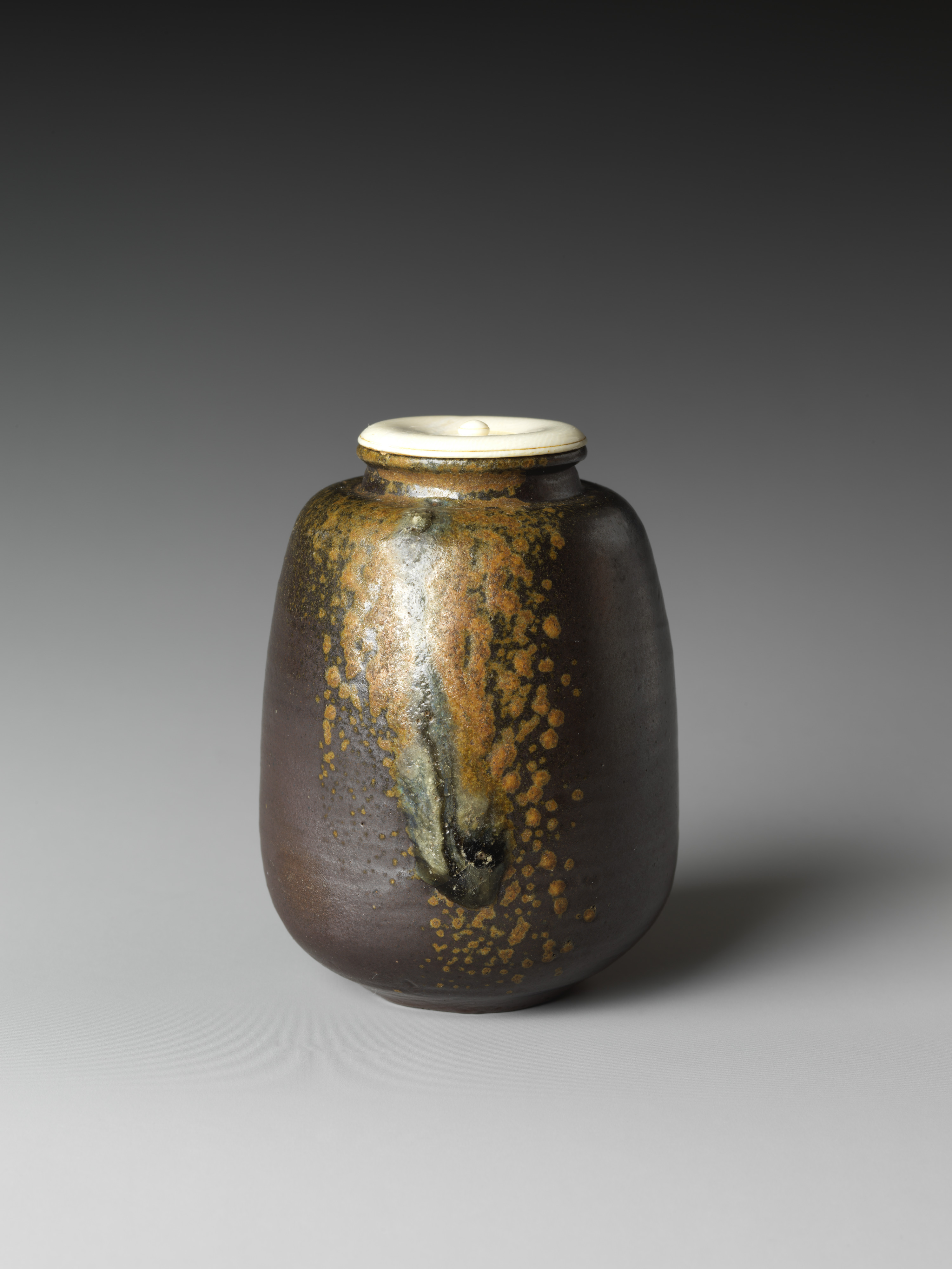 A seventeenth-century Bizen tea caddy in the Metropolitan Museum of Art, its dark brown body crossed by a single run of ash glaze and speckled amber, with a pale ivory lid