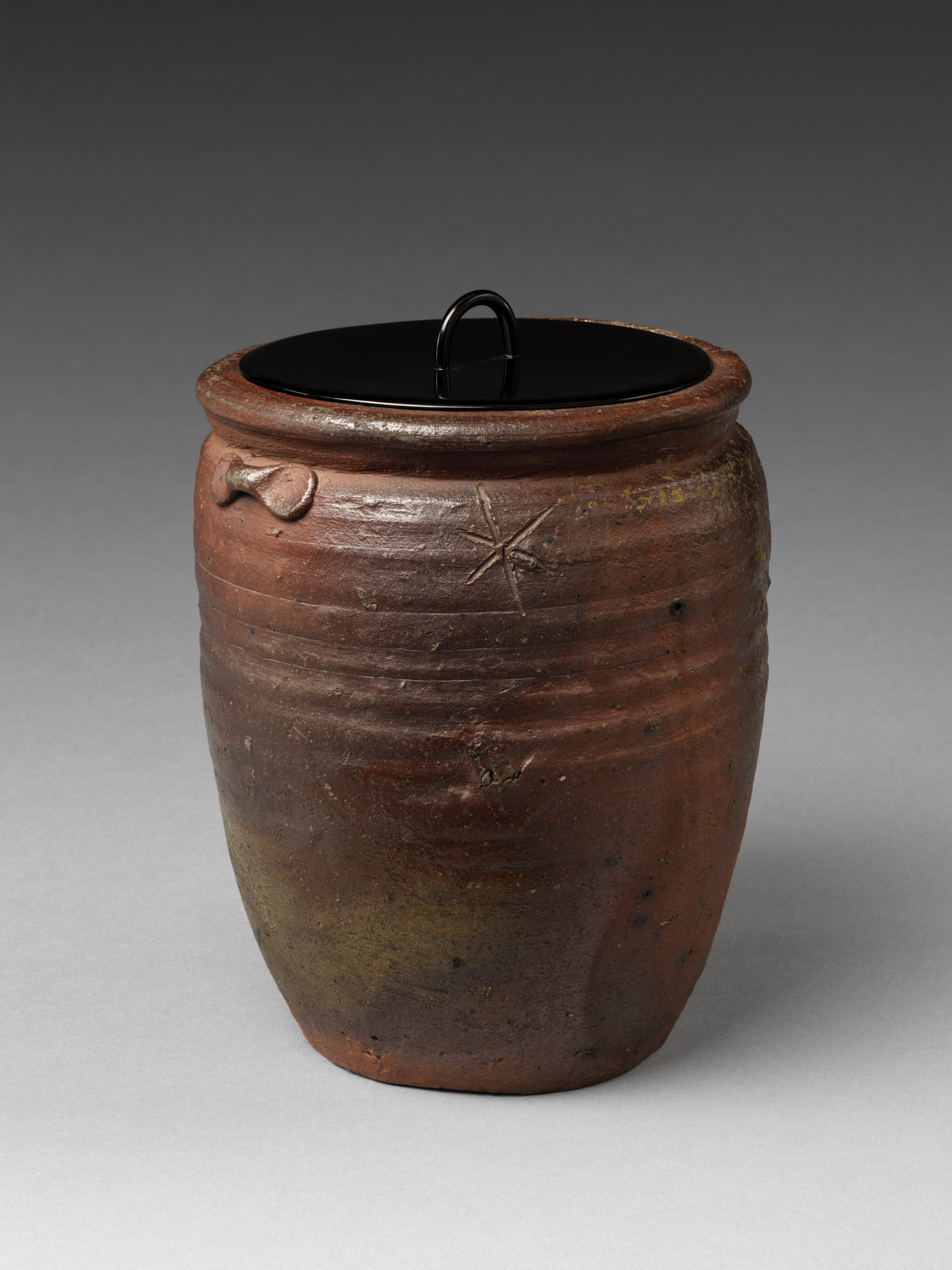 A late sixteenth-century Bizen freshwater jar in the Metropolitan Museum of Art, reddish-brown and lightly ridged, with an incised star-shaped mark of three crossed strokes and an applied lug at the shoulder, under a black-lacquered wooden lid with a metal ring