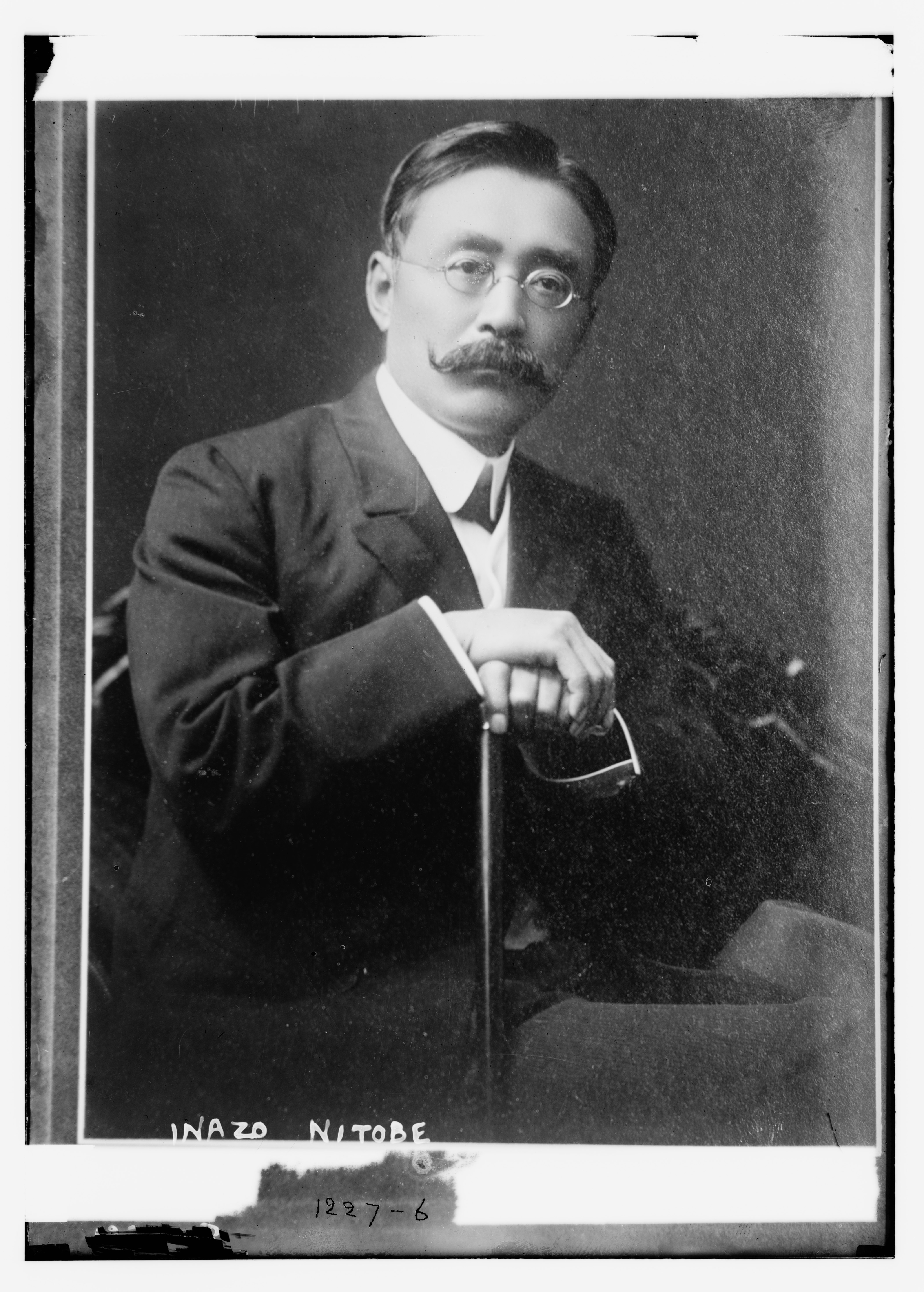 A black-and-white portrait photograph of Inazo Nitobe in a Western suit and round wire-rim glasses, resting a hand on a walking cane