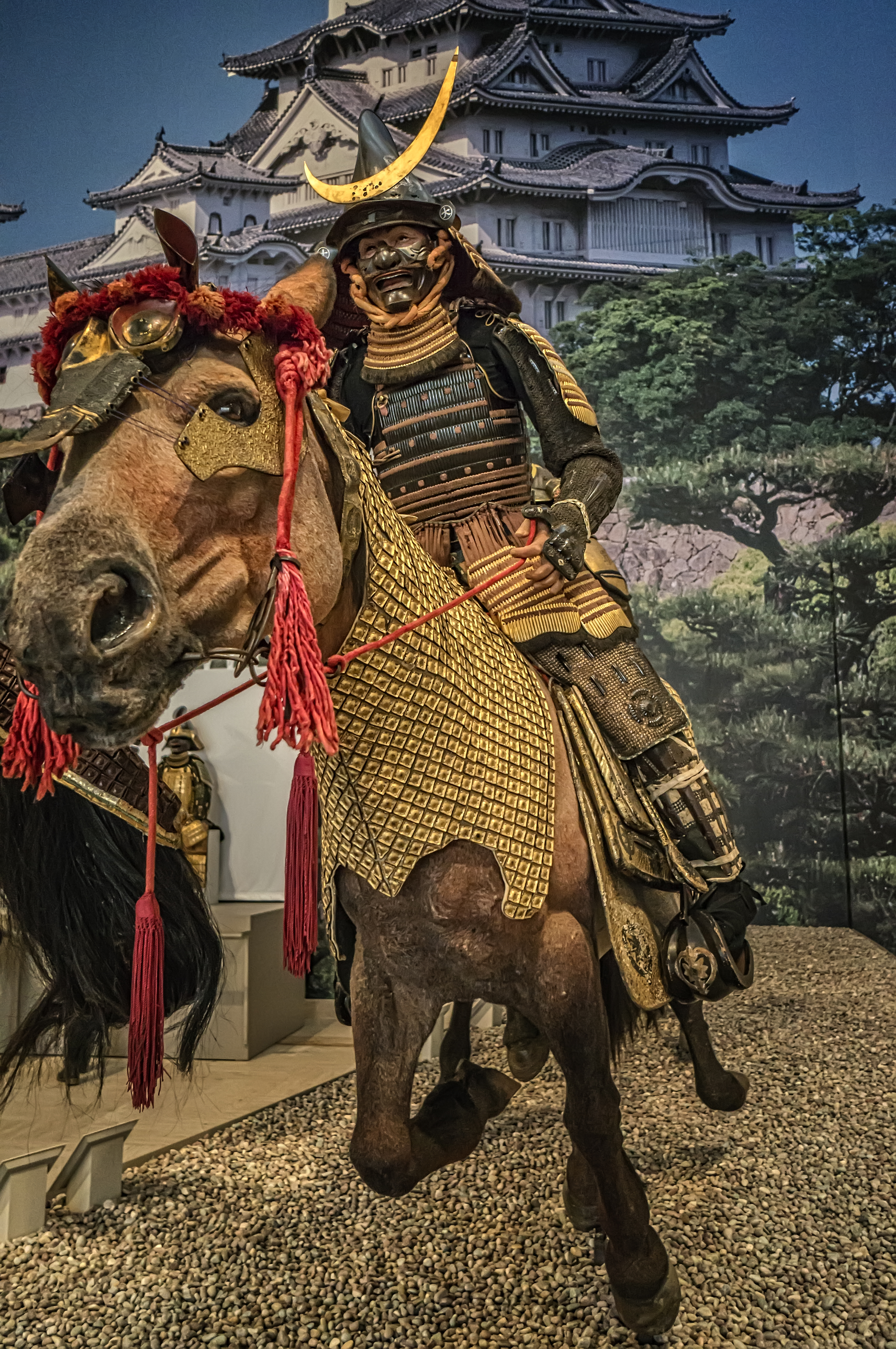 A museum display of a mounted samurai in gold-laced Tachido armor and a crescent-moon helmet crest, staged before a painted backdrop of a Japanese castle