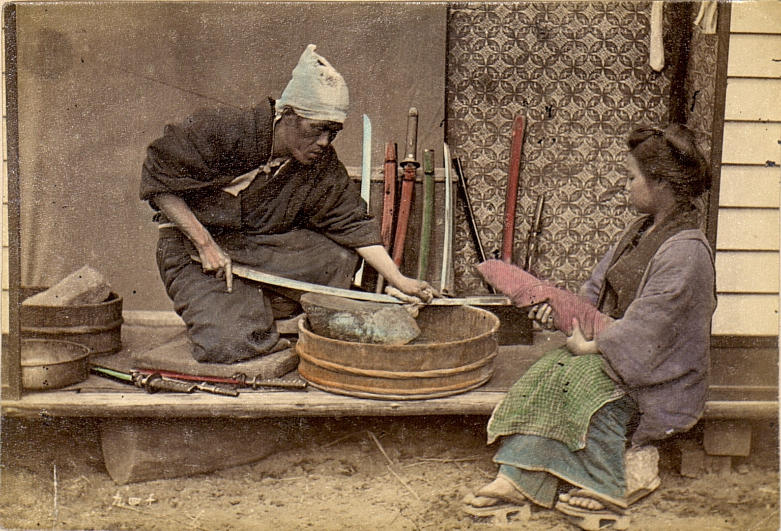 A hand-tinted historical photograph of a sword sharpener kneeling to hone a blade over a large wooden whetstone basin, with several sheathed swords standing beside him