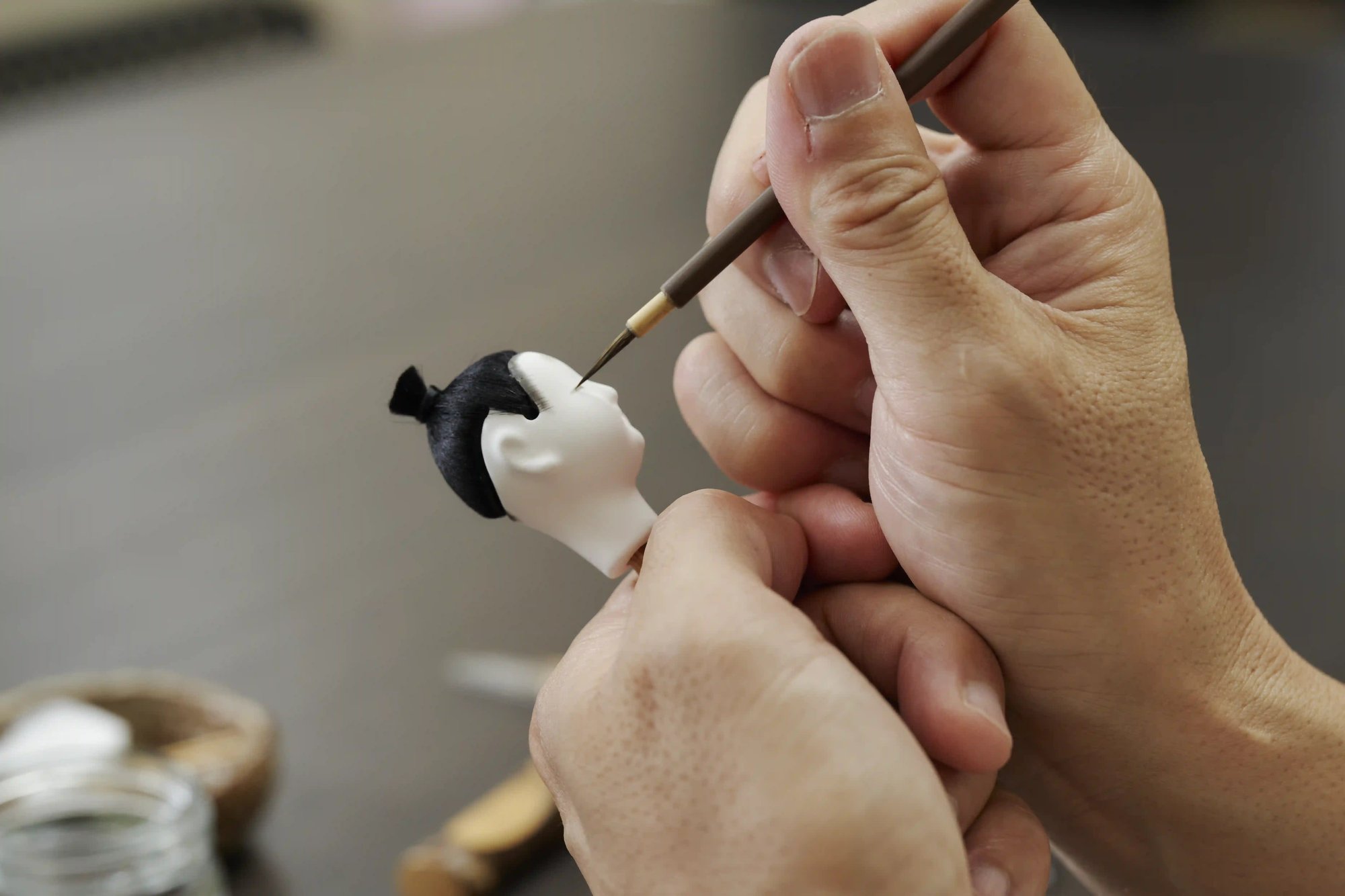 A craftsman's hands holding a hina doll head steady while a fine brush paints its face