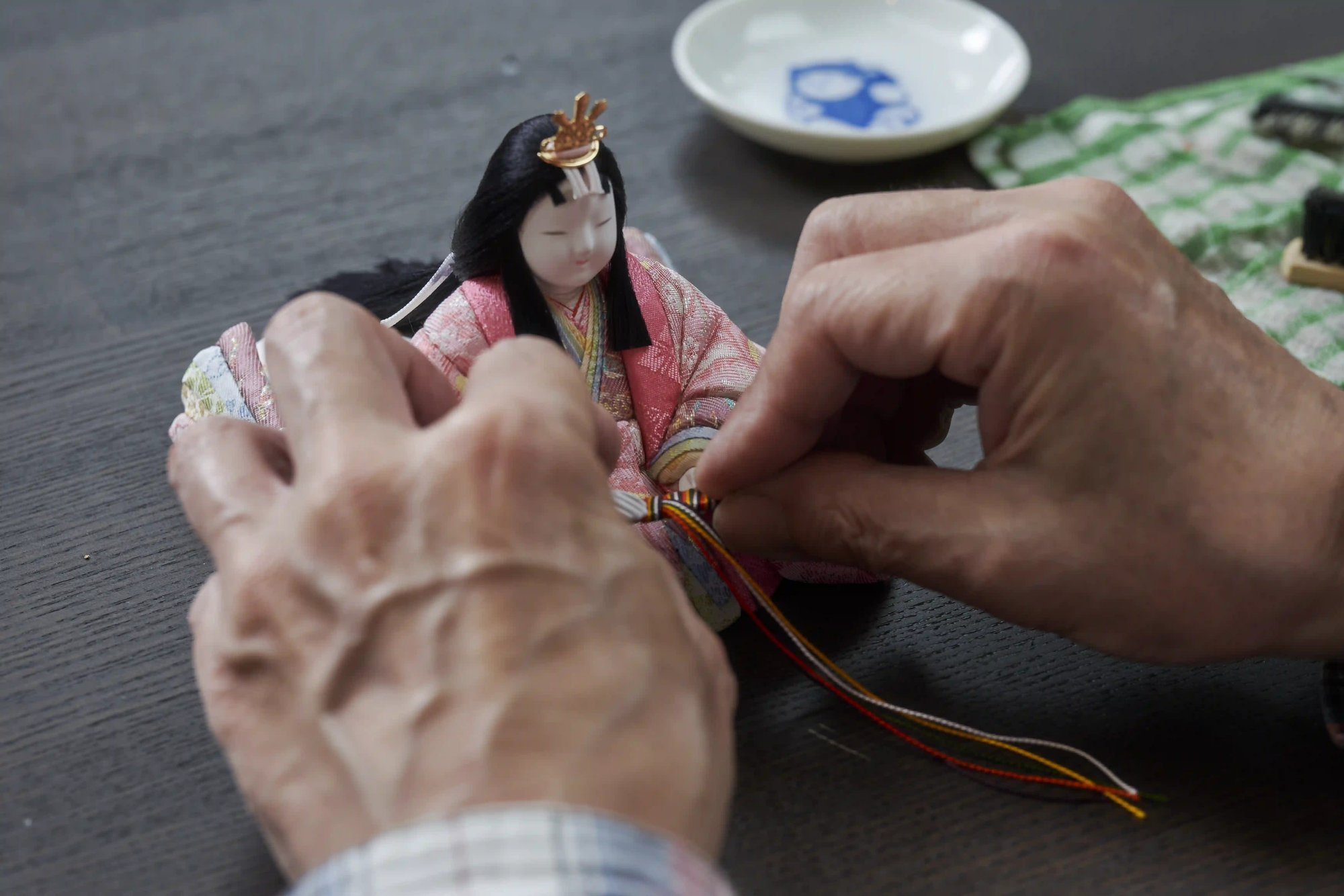 A craftsman's hands tying the cords of a costume on a small hina doll wearing a gold crown, at a workbench