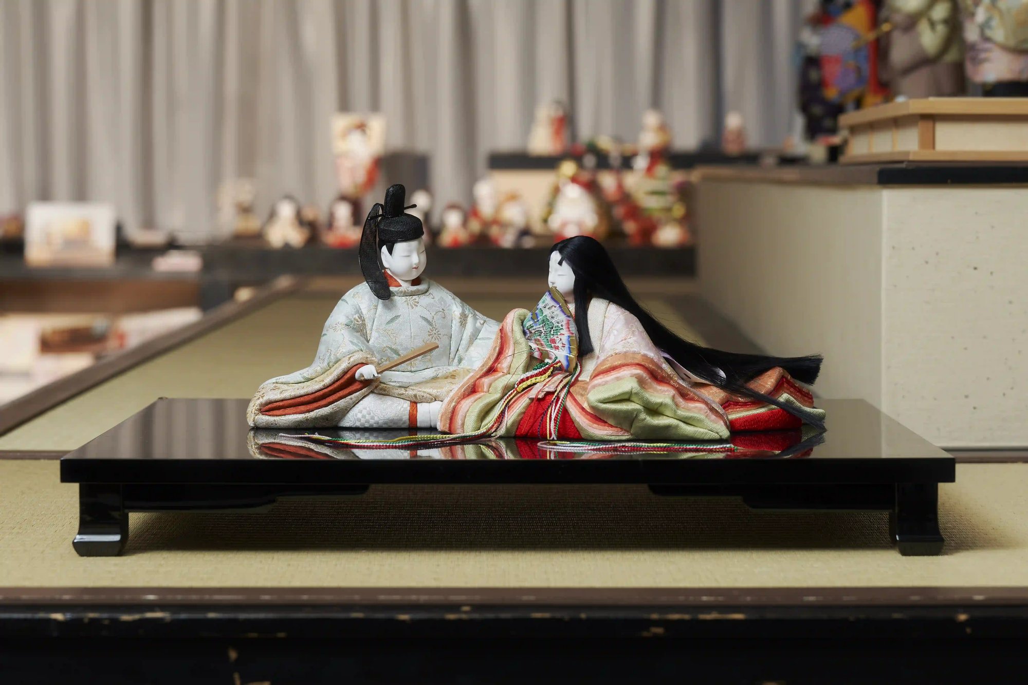 A male and a female hina doll in layered court robes, seated facing each other on a low black lacquer stand, with more dolls on display behind them