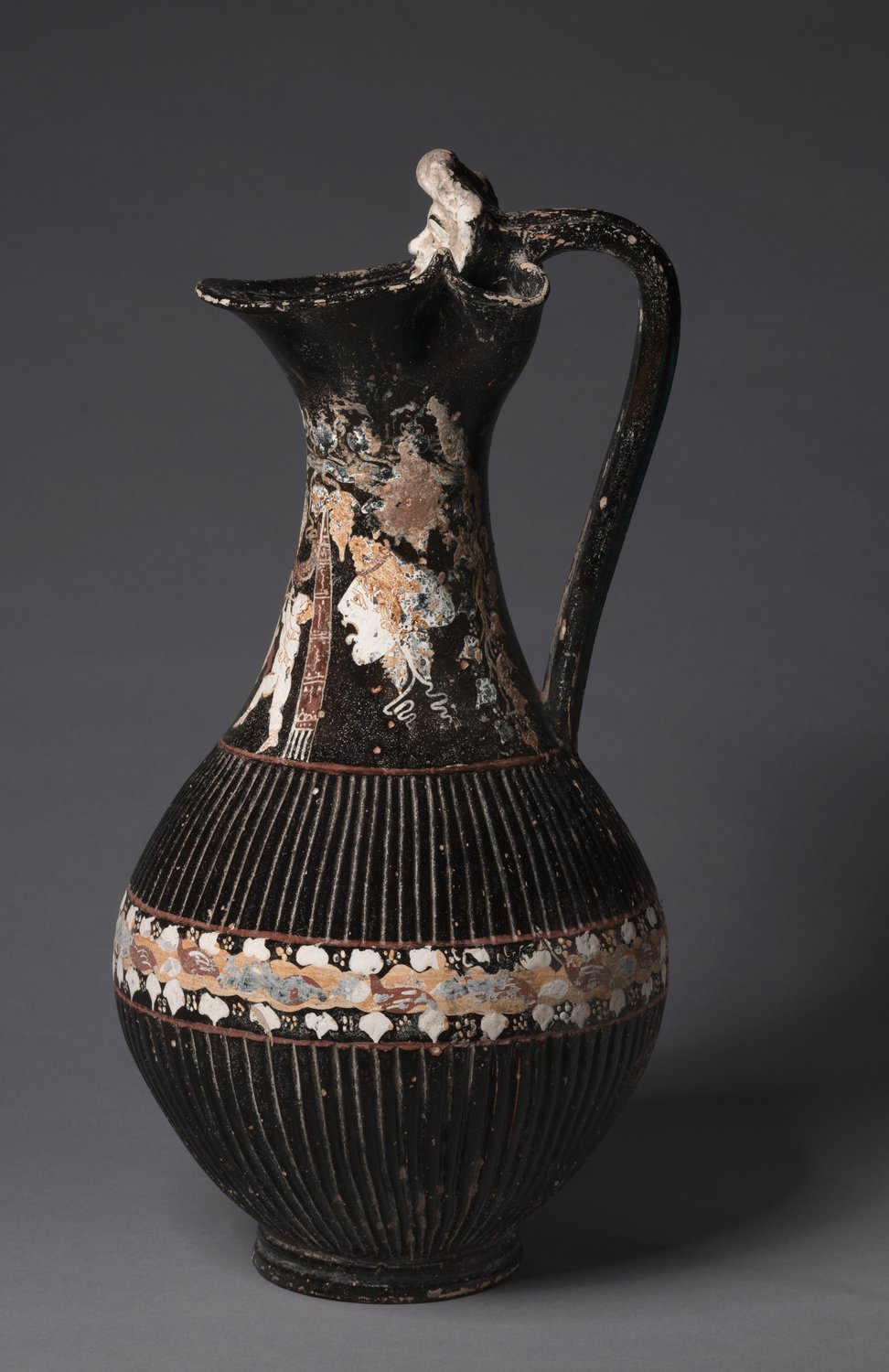 A tall black South Italian wine jug of about 300 BCE with a small moulded theatre mask at the top of its handle and painted figures on its neck