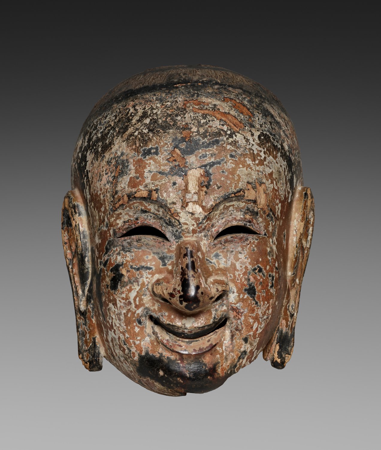 A worn wooden gigaku mask of a smiling young man with large ears, its lacquer and paint partly lost