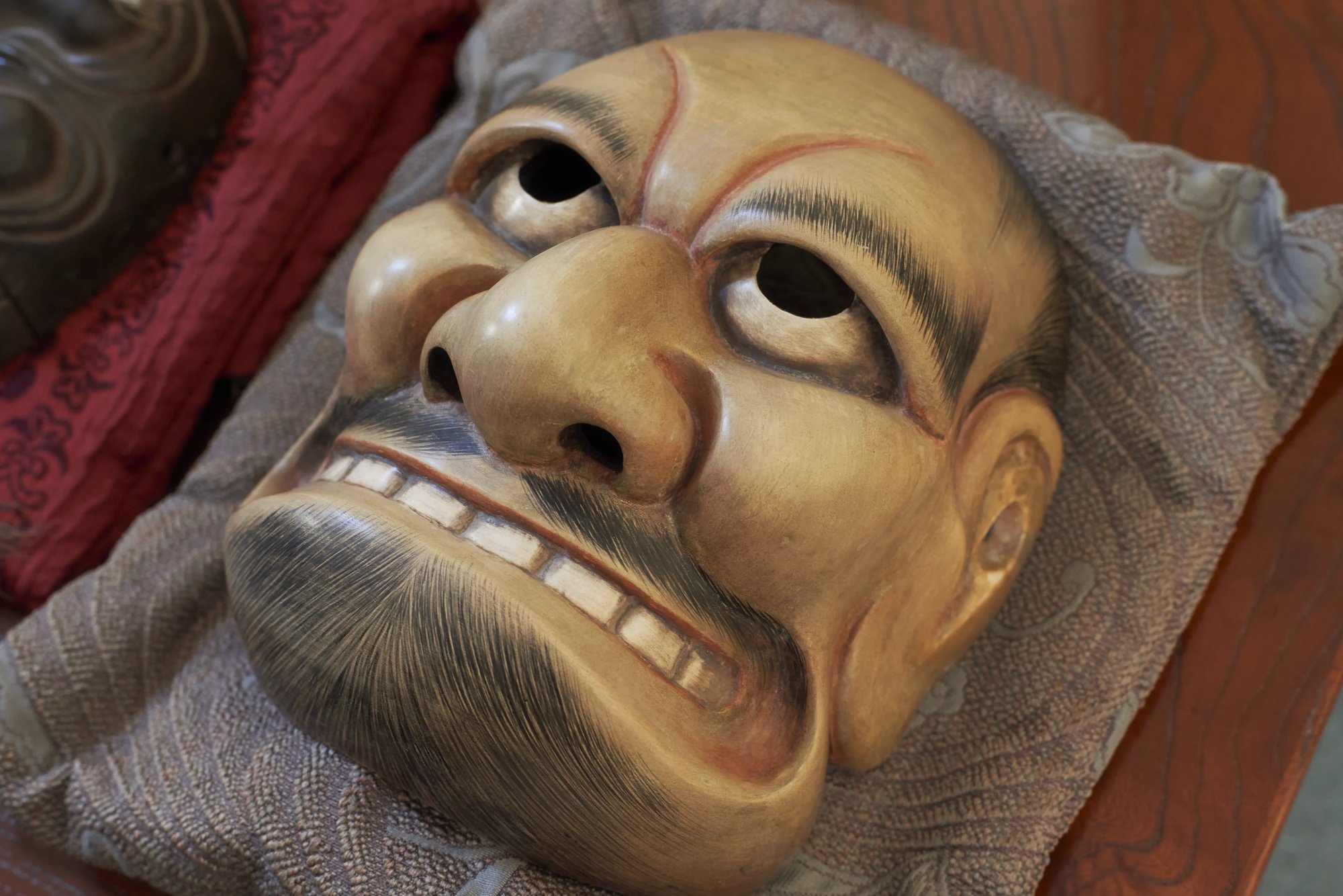 A kyogen mask tilted toward the camera, showing its eye holes, heavy brows, teeth and worn varnish