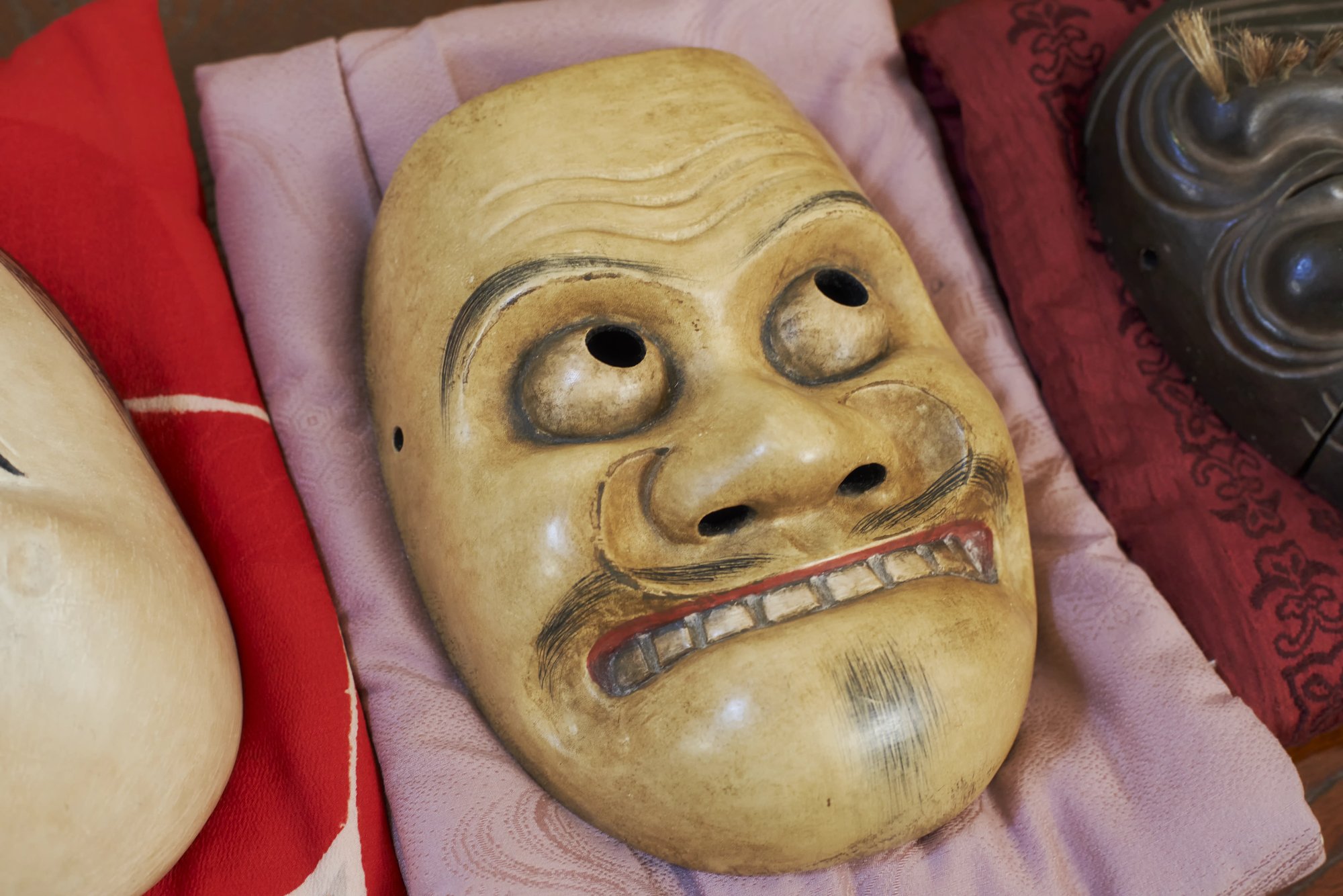 Kyogen masks laid out on folded cloths, a pale mask in the foreground and a dark one behind it