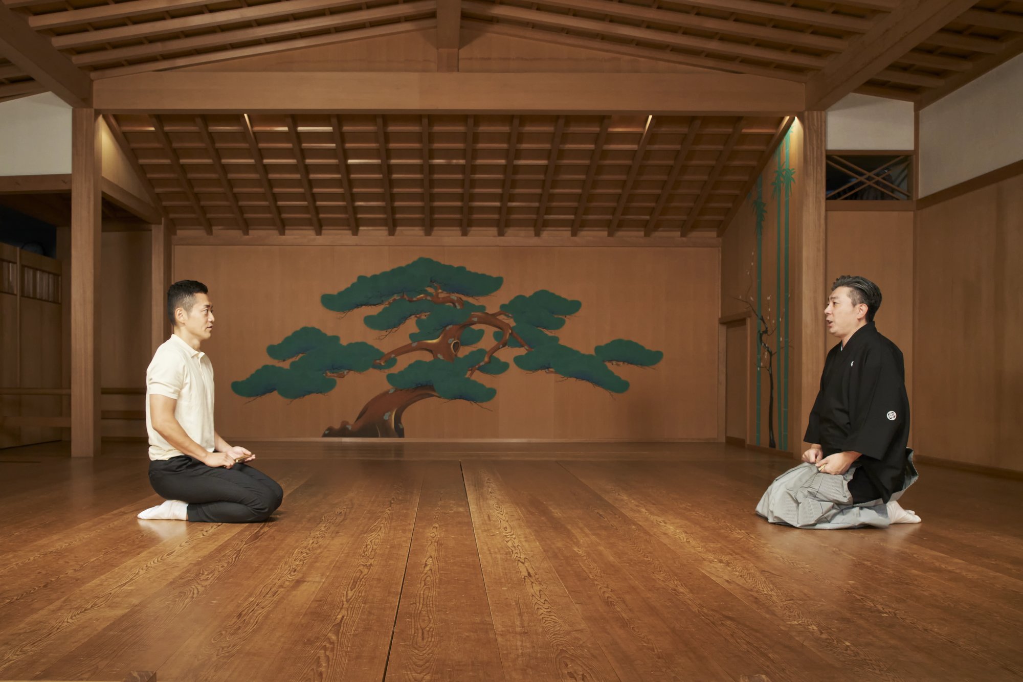 Two kyogen performers kneeling opposite each other on a noh stage, with a painted pine on the back wall