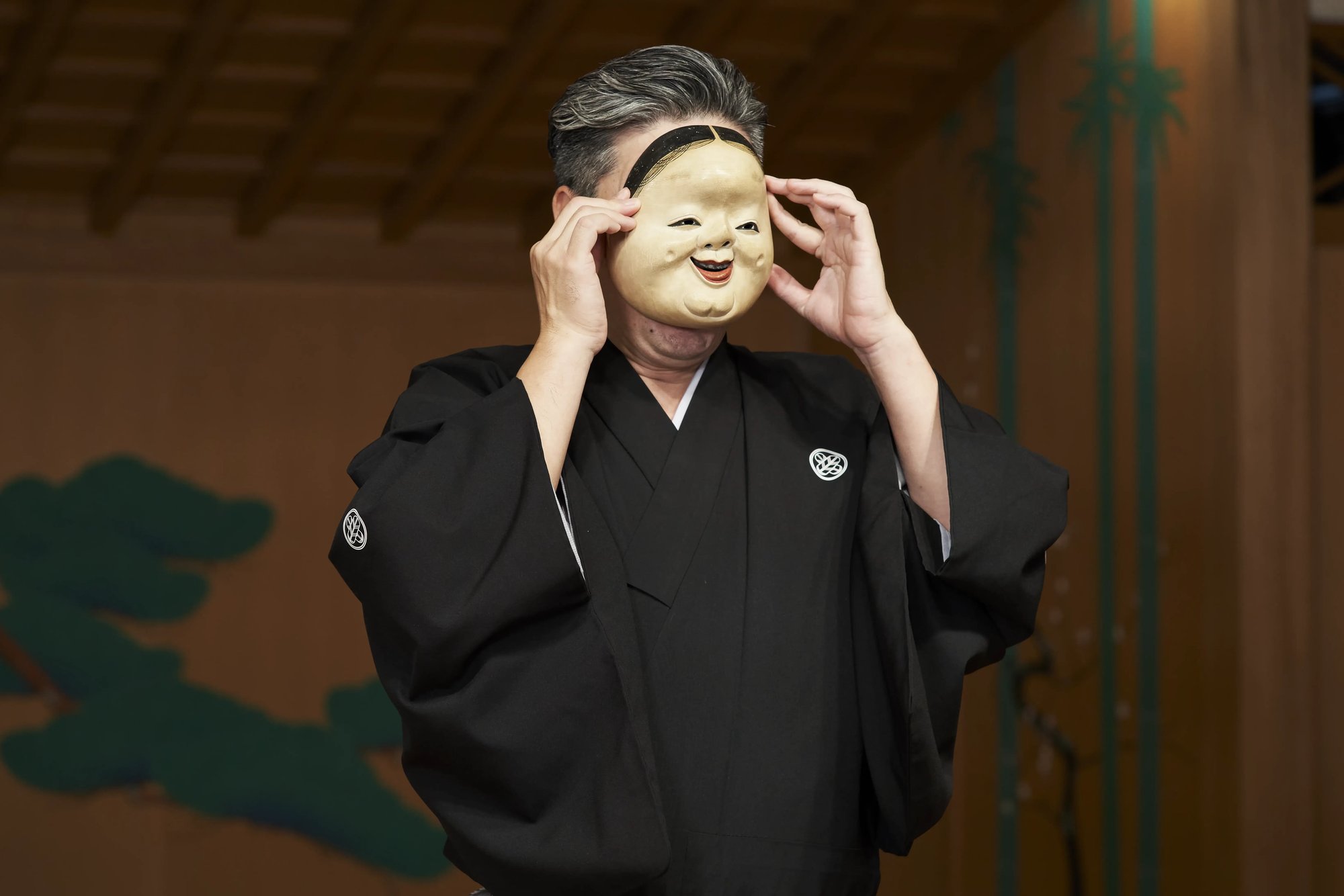 A kyogen performer in a black formal robe lifting a smiling mask toward his face on a noh stage