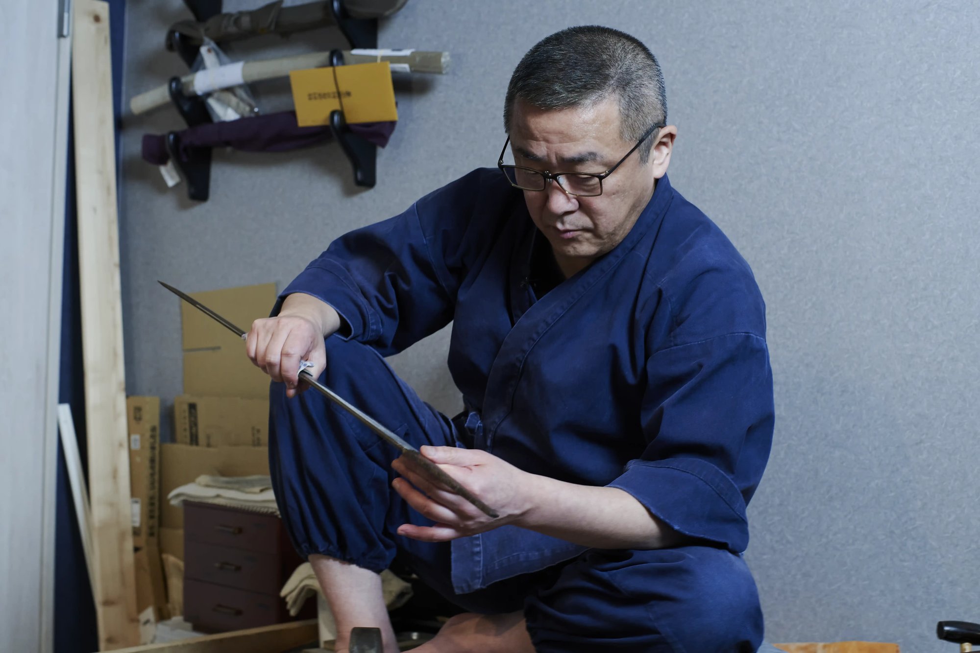 A sword polisher in an indigo work jacket sitting on the floor and holding a bare blade up to examine it