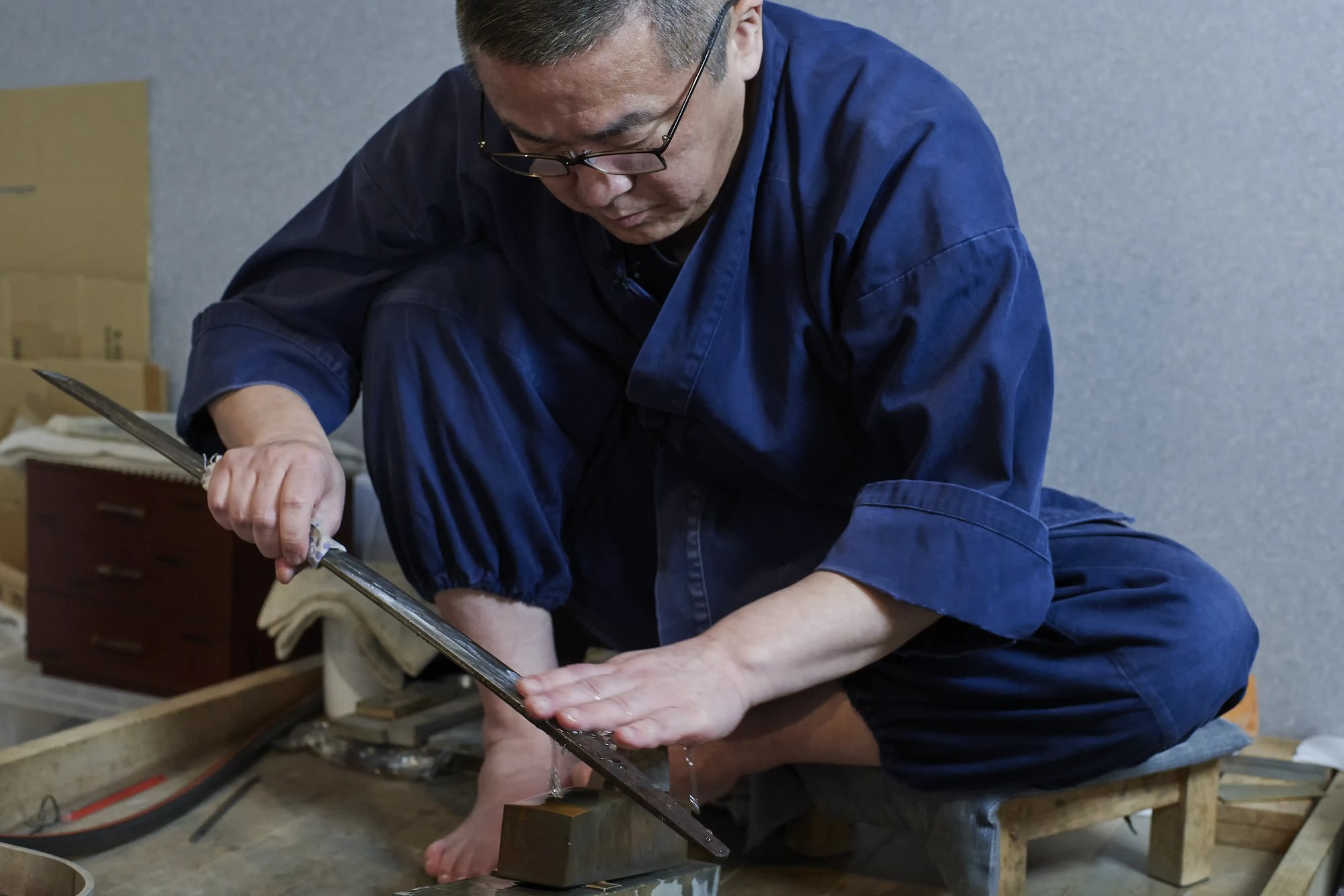 A sword polisher seated on the floor, working a long blade against a stone with both hands