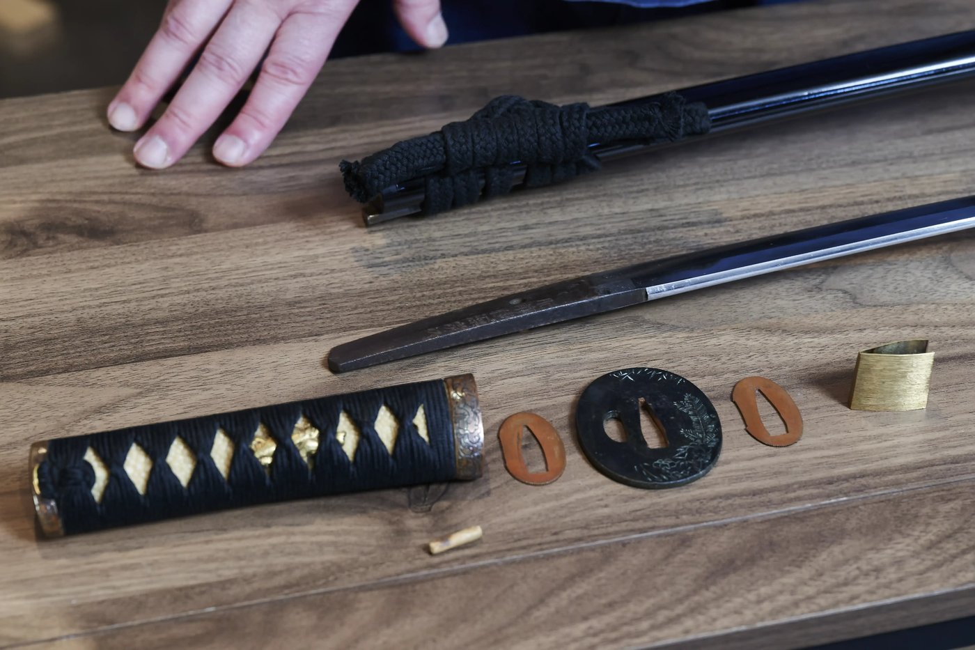 A sword taken apart on a wooden table: the wrapped handle, guard, spacers and collar laid out in a row beside the bare blade and its scabbard