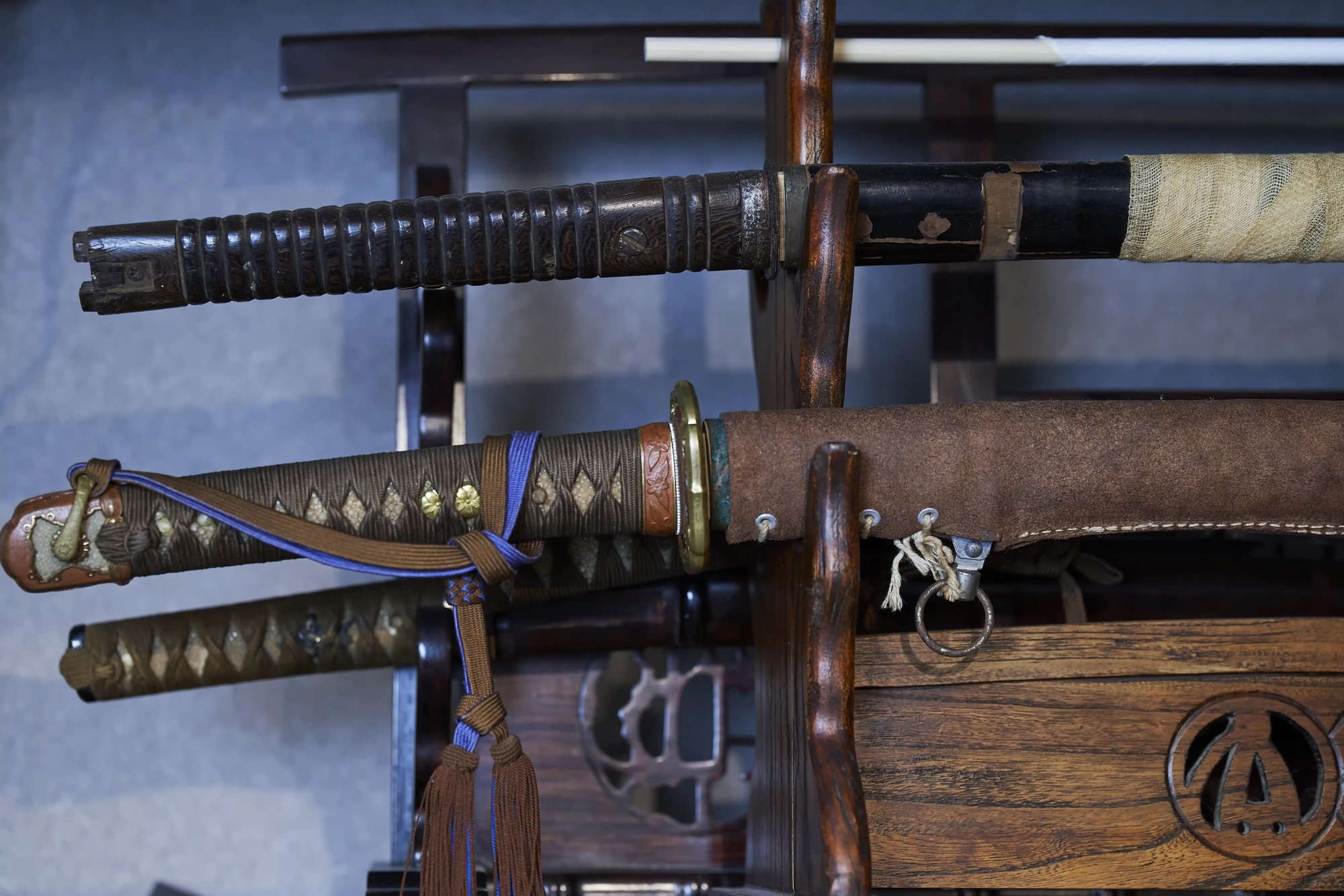 Swords in military-style mountings resting on a wooden stand: a ribbed grip and cloth-wrapped scabbard above, a braid-wrapped grip with a tassel and a leather-covered scabbard below