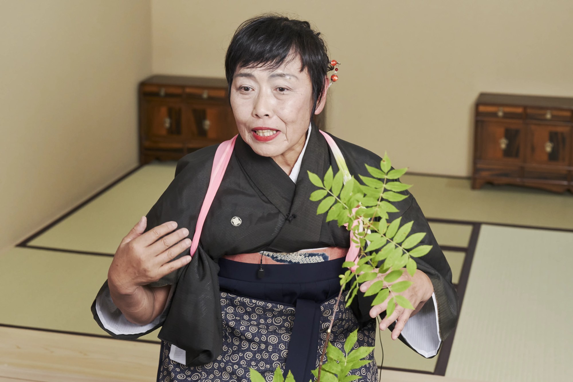 An ikebana teacher in a dark kimono explaining with a raised hand, leafy branches in front of her