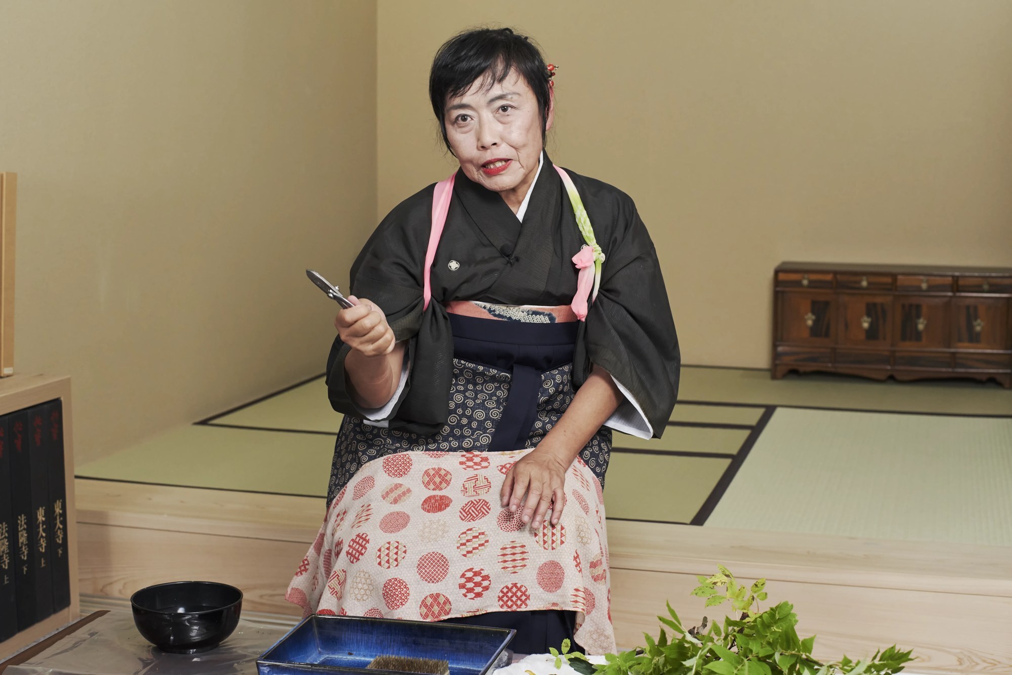 An ikebana teacher in a dark kimono looking straight at the camera, shears in hand, a blue basin before her