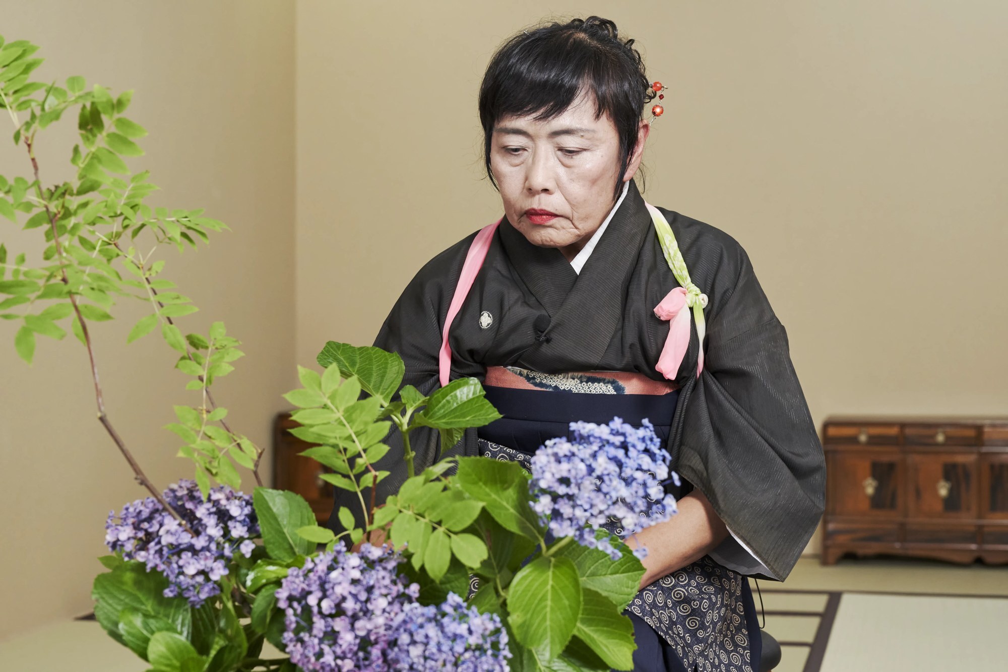 A kimono-clad ikebana teacher seated behind a driftwood arrangement of hydrangea