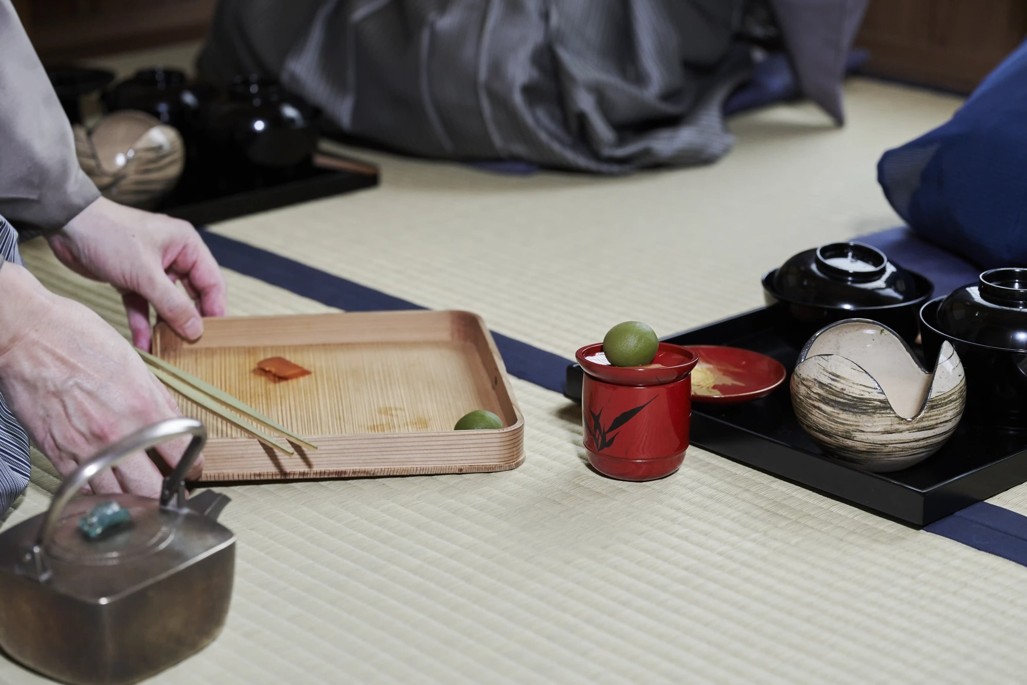 A tray of kaiseki dishes and a red lacquer cup on tatami, beside a kettle and a wooden tray
