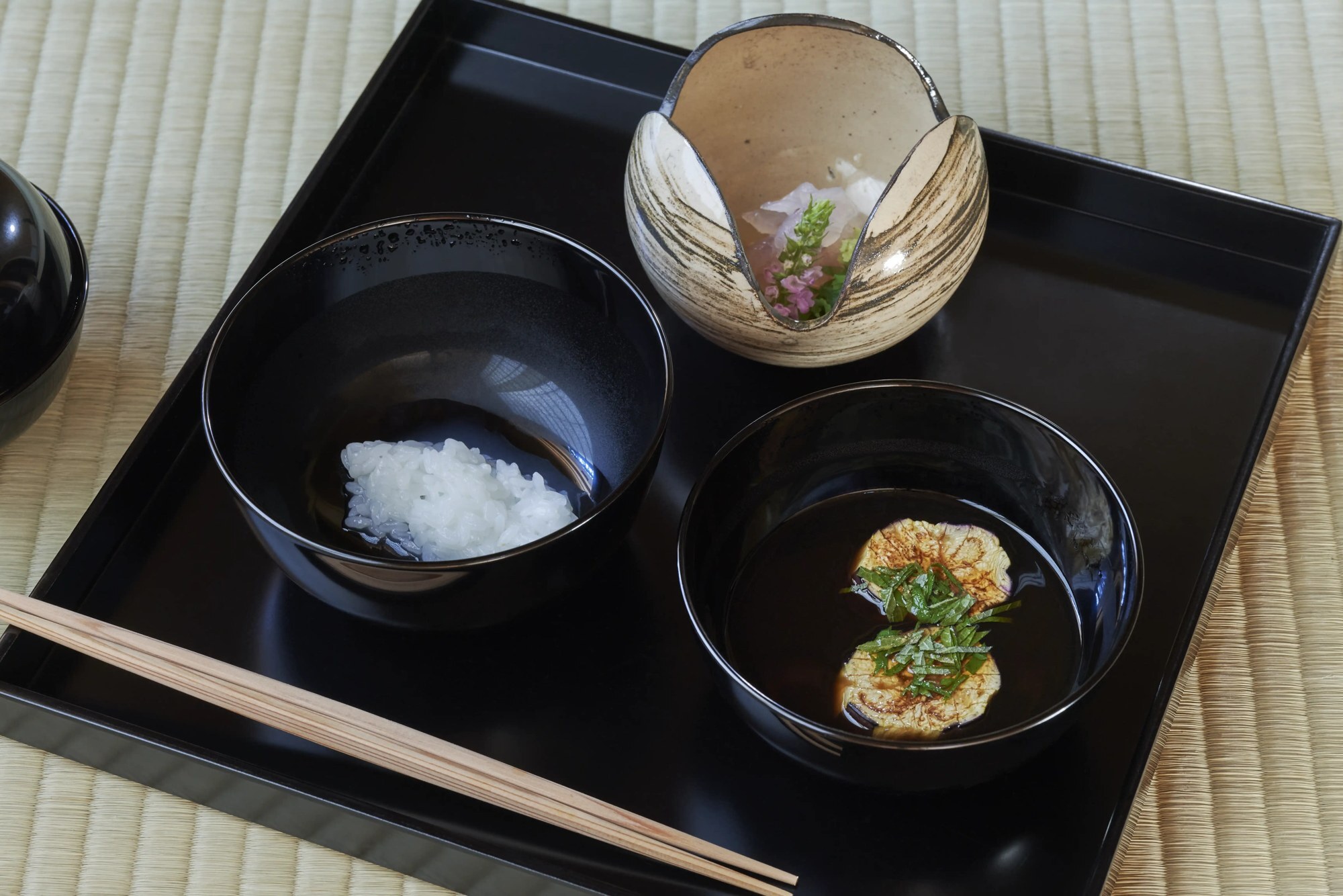 A black lacquer tray with a rice bowl, a soup bowl, a ceramic dish and chopsticks on tatami