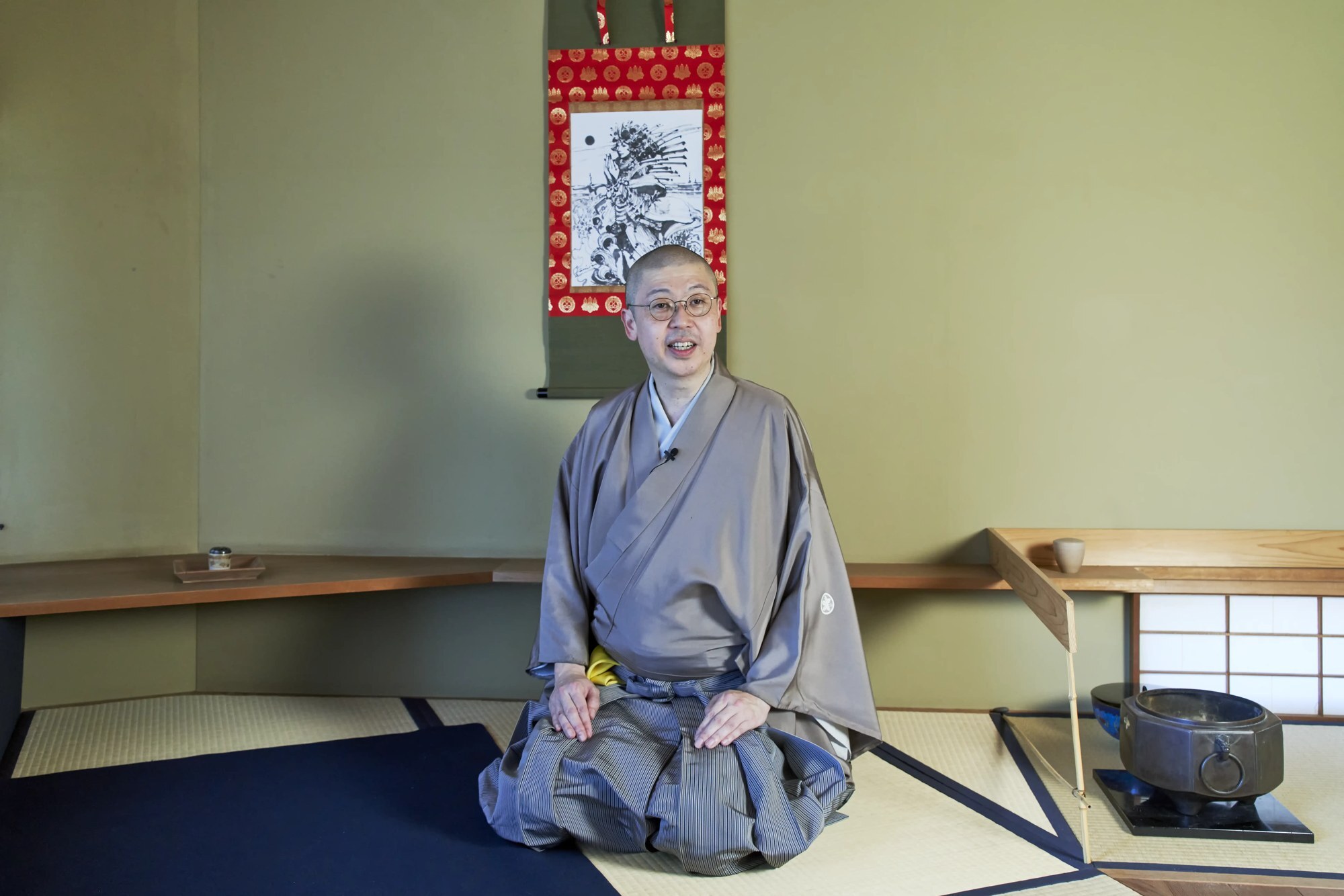 A tea host in a grey kimono seated on tatami in a tea room beside an iron kettle