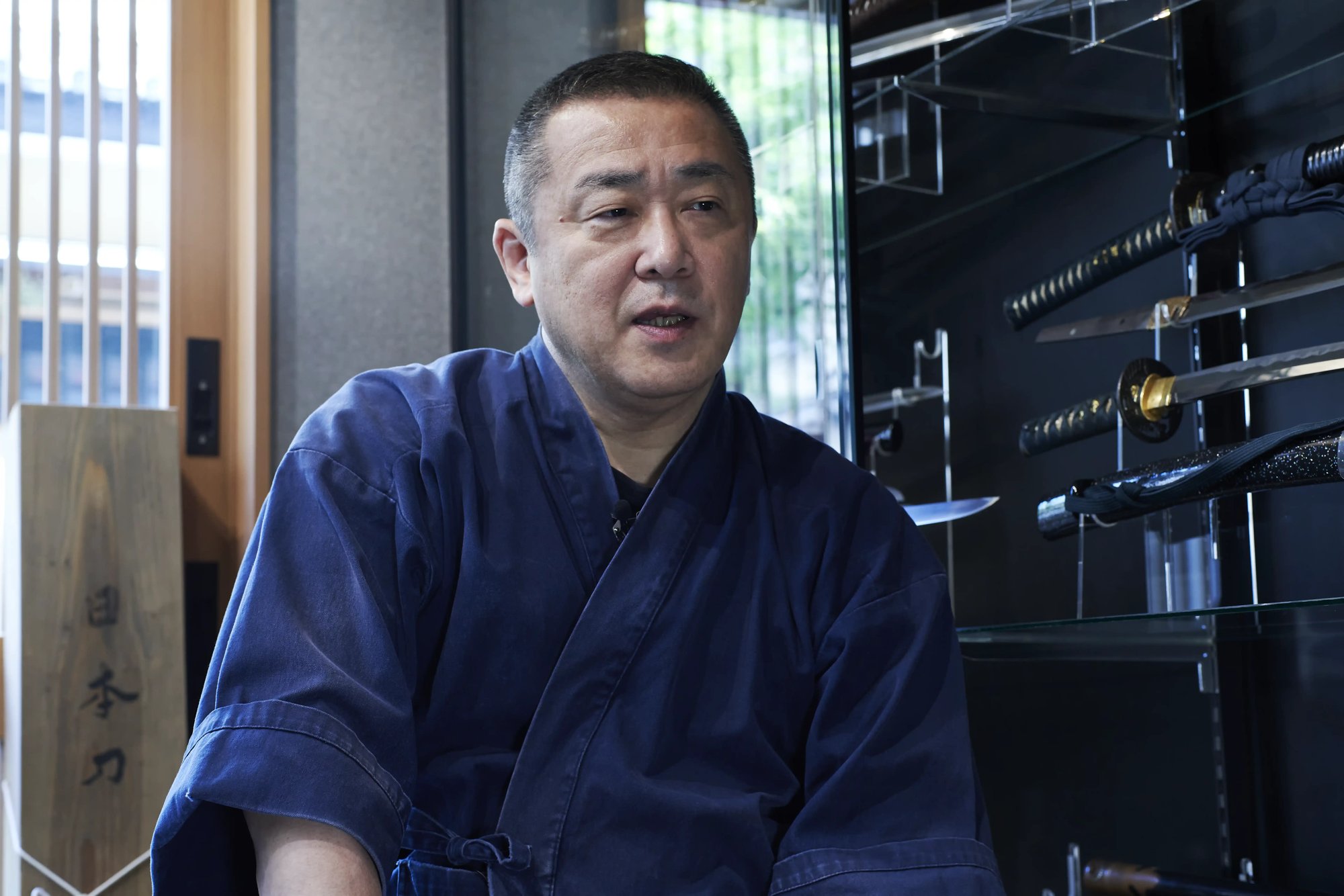 A sword polisher in an indigo work jacket speaking in his workshop, with glass cabinets of swords and a sign reading Nihonto behind him
