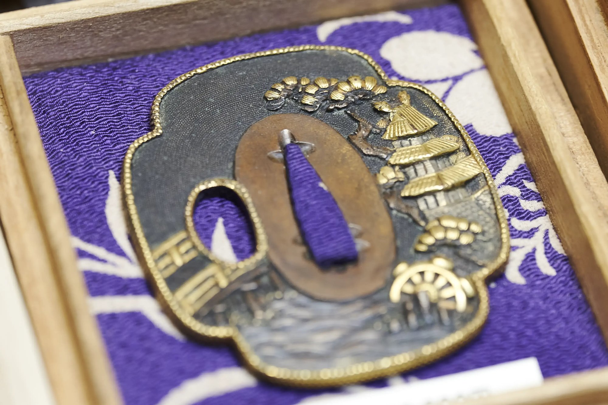 A sword guard with a gilt landscape design, framed on purple cloth in a wooden case