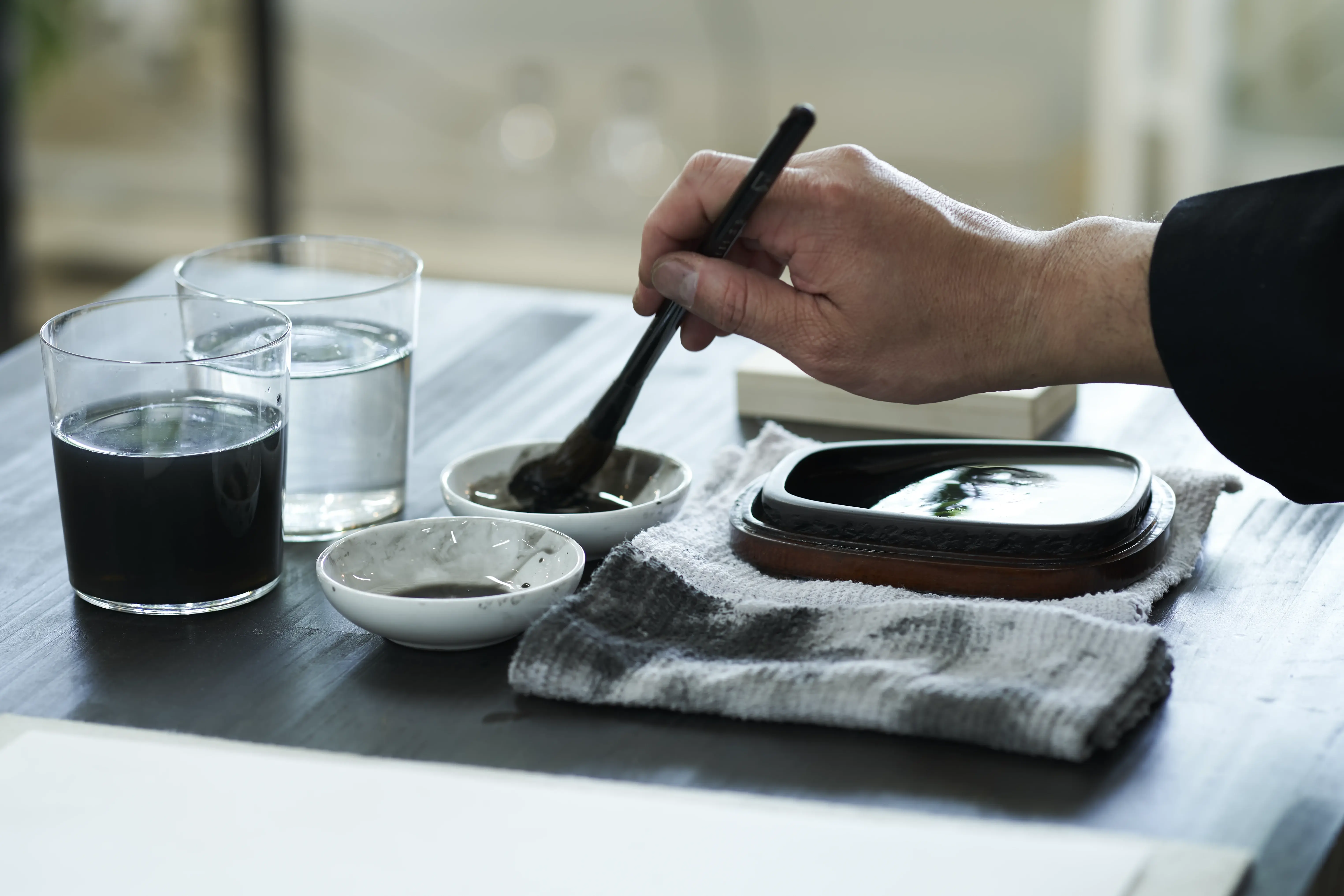 A hand grinding an inkstick on a stone inkstone, with two small dishes of ink and two glasses of water on the work table