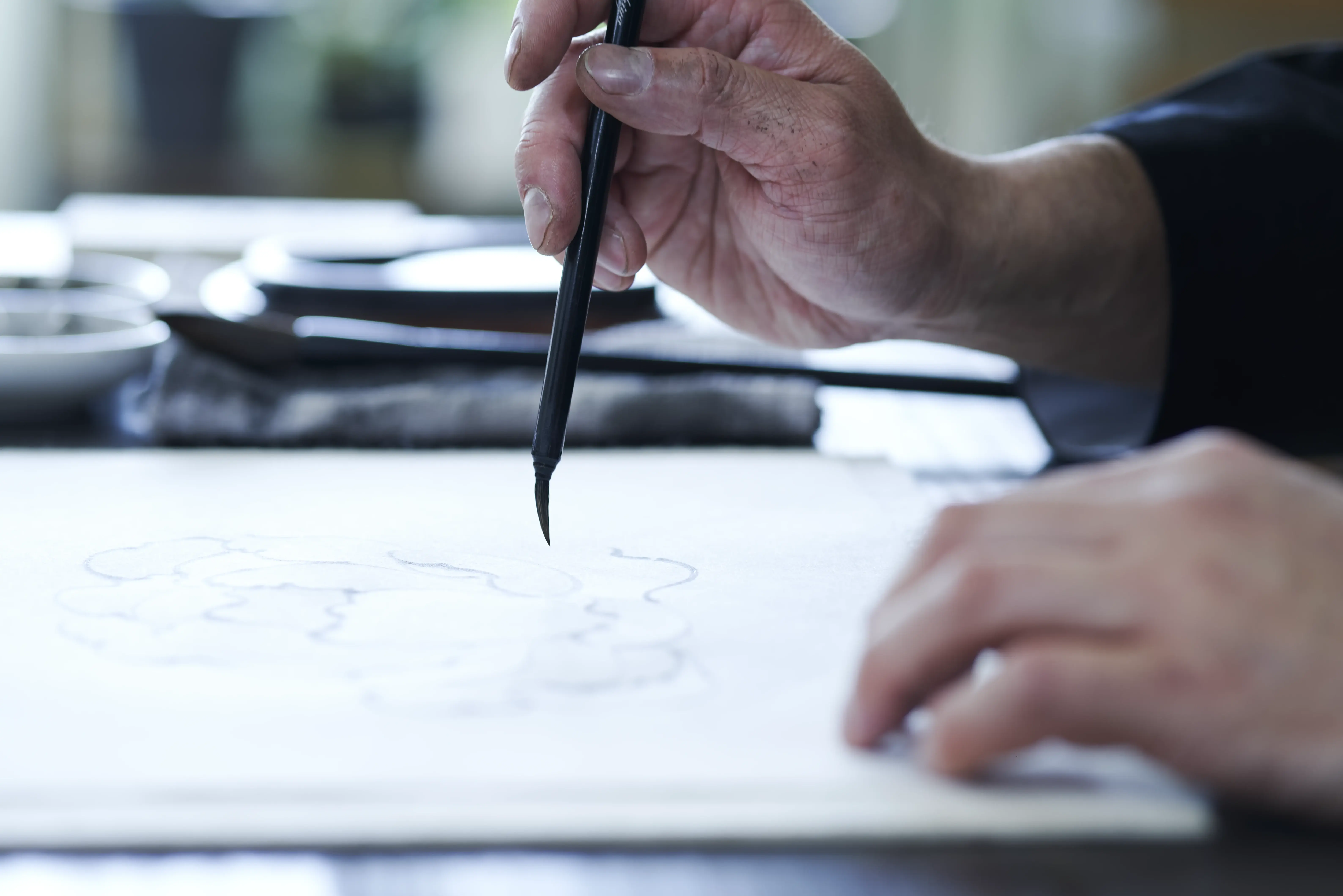 A brush touching washi at the start of a stroke, an outline of a composition already drawn on the sheet beneath it
