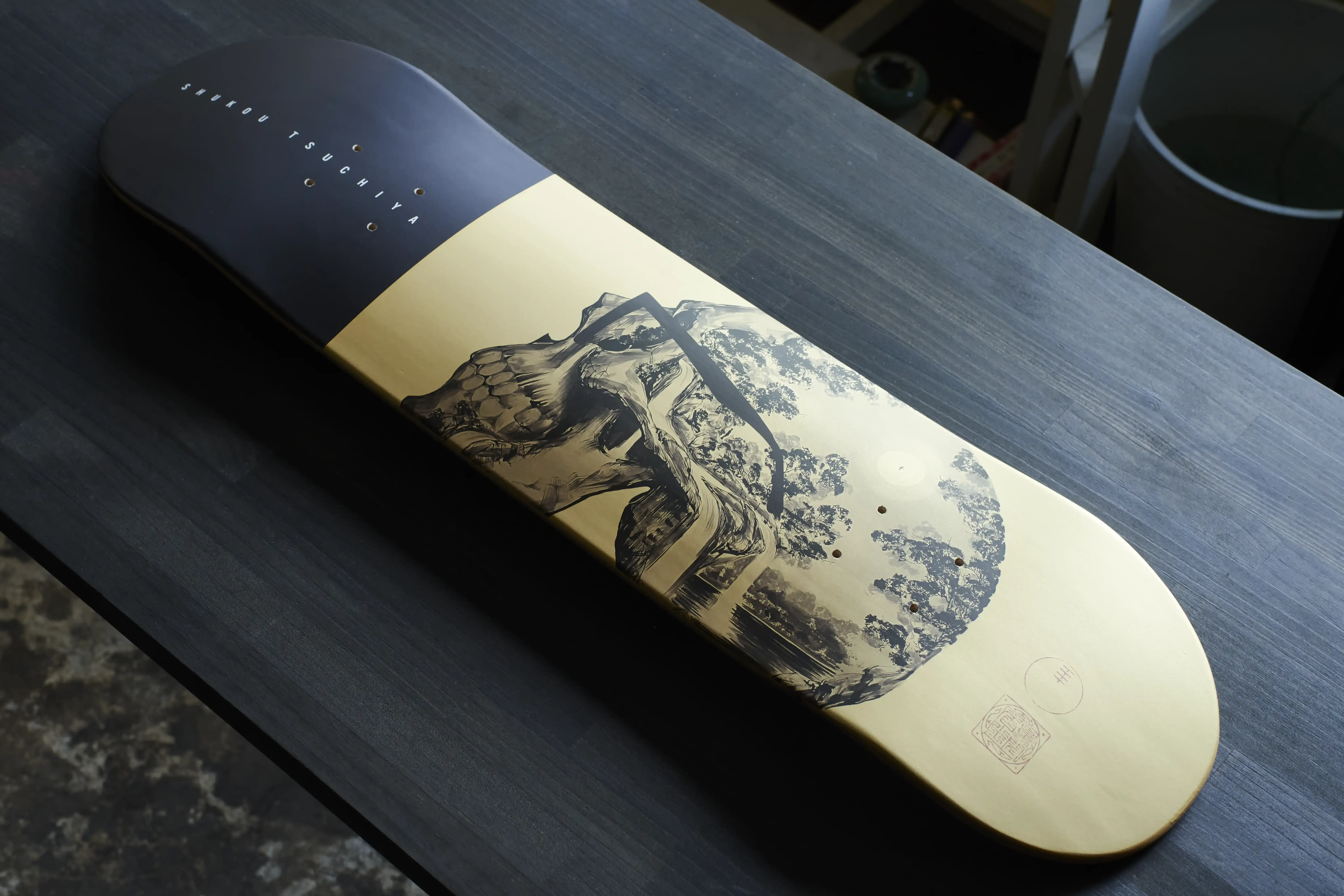 A skateboard deck printed with an ink landscape, resting on a dark workbench