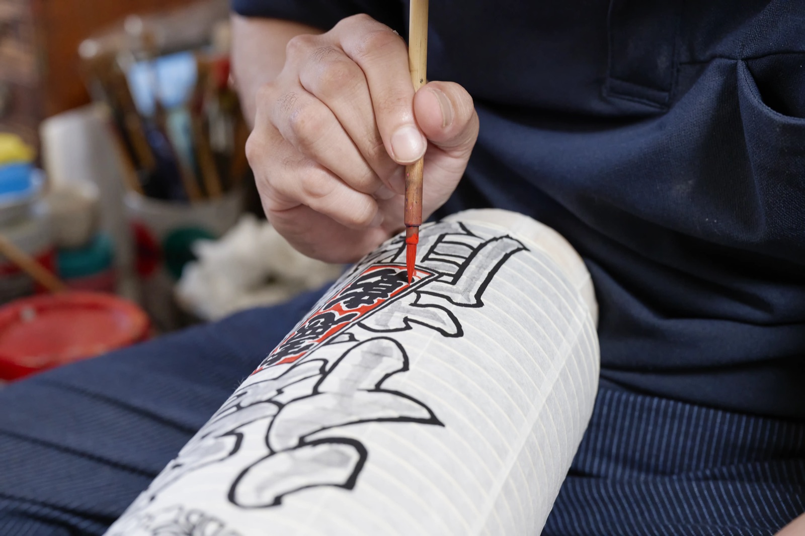 A brush held against the washi surface of a chochin, the evenly spaced bamboo ribs visible beneath the paper