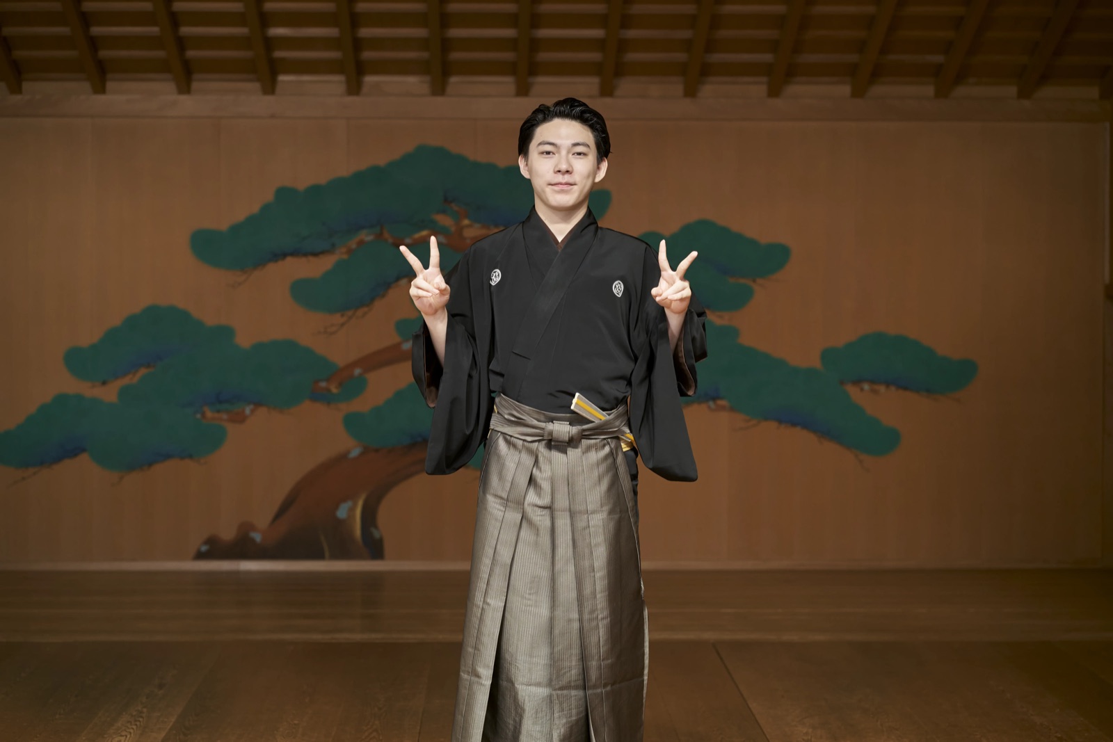 A kyogen performer standing on a bare hinoki cypress noh stage, the painted pine of the kagami-ita filling the wall behind him