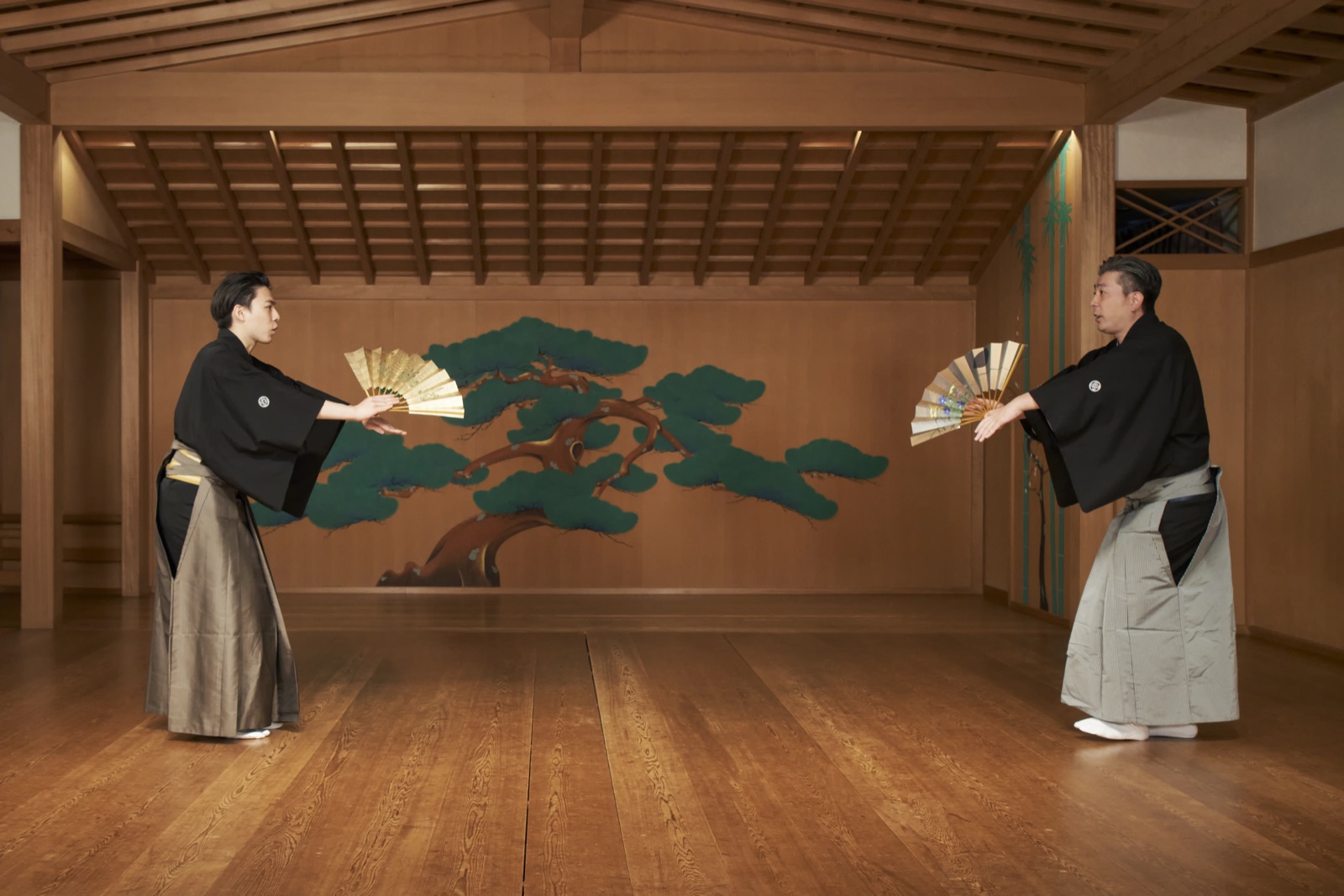 Two kyogen performers facing each other across the noh stage, each holding an open folding fan