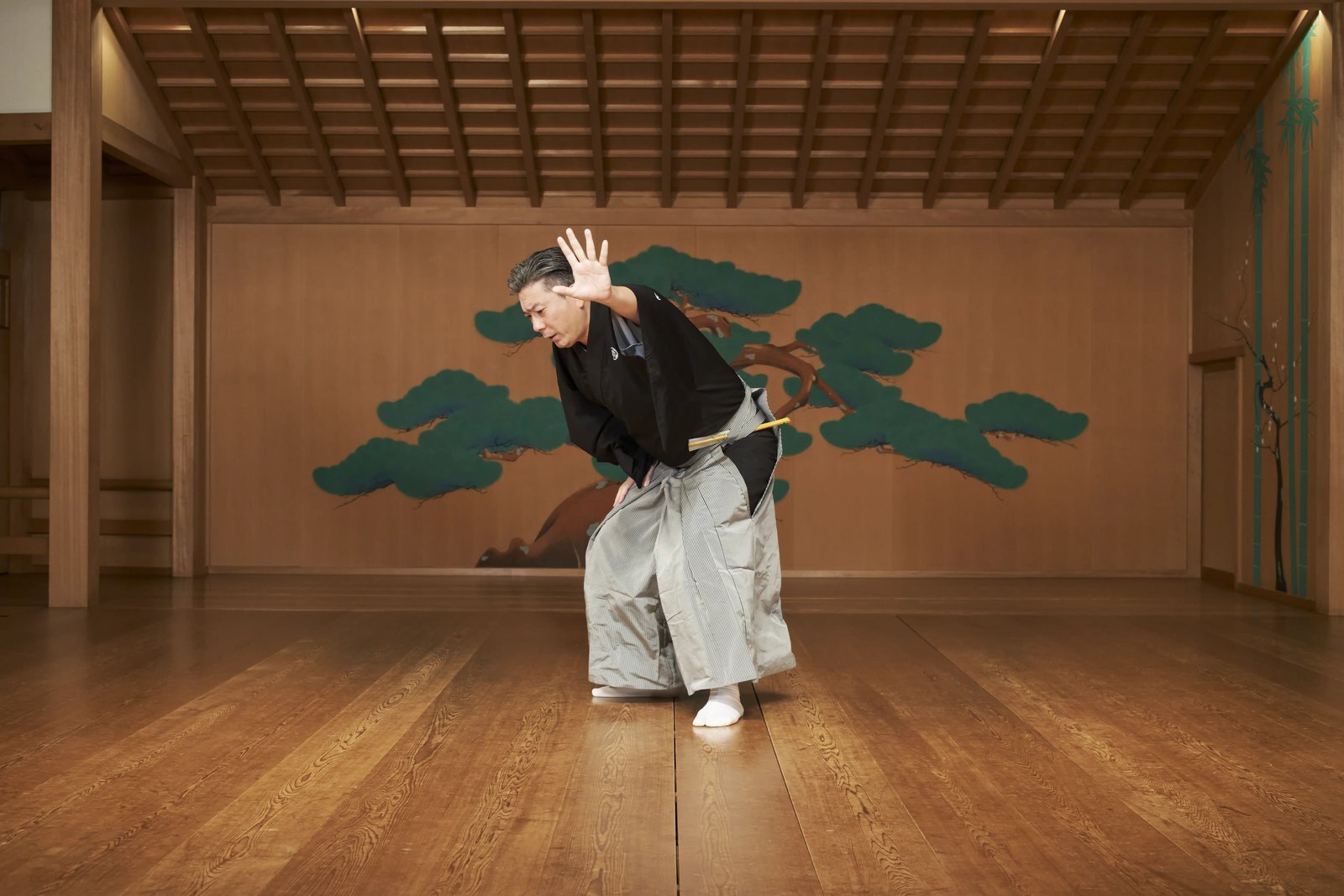 A kyogen performer bowing on the stage, the hall's timber ceiling, corner pillar and the pine painted across the back wall all in view