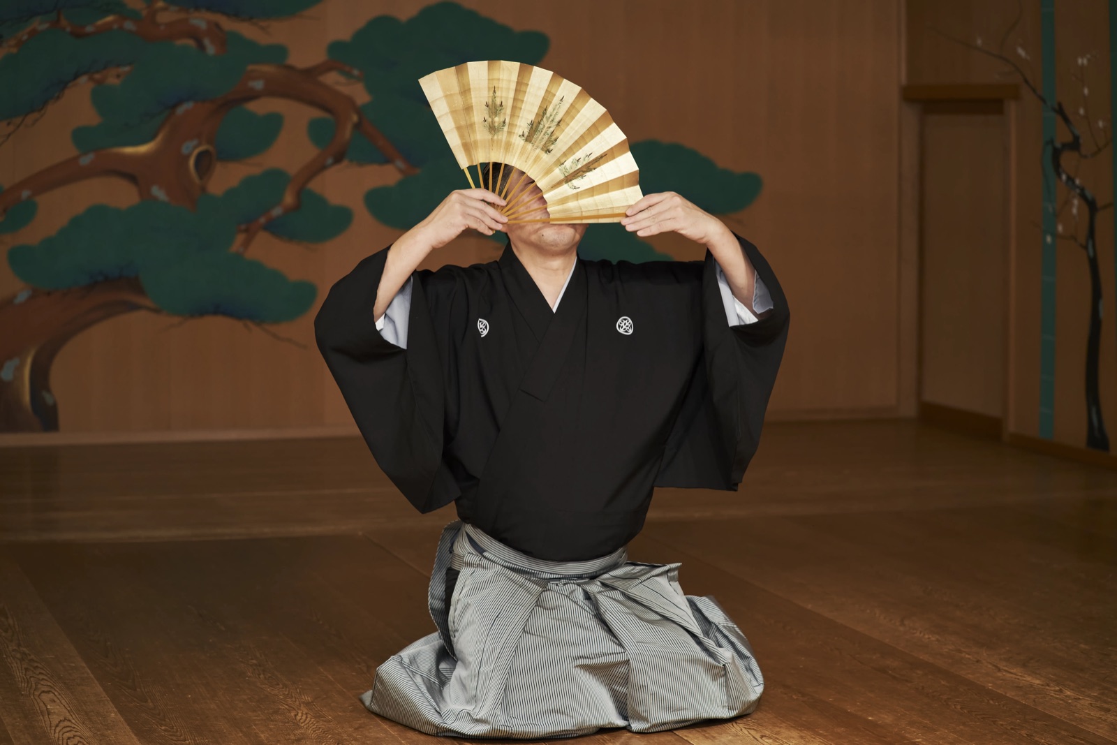 A kyogen performer kneeling on the stage boards with a folding fan opened in front of his face