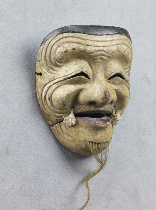 A noh mask of the Okina type seen from a three-quarter angle, its chin carved separately and tied on with cord