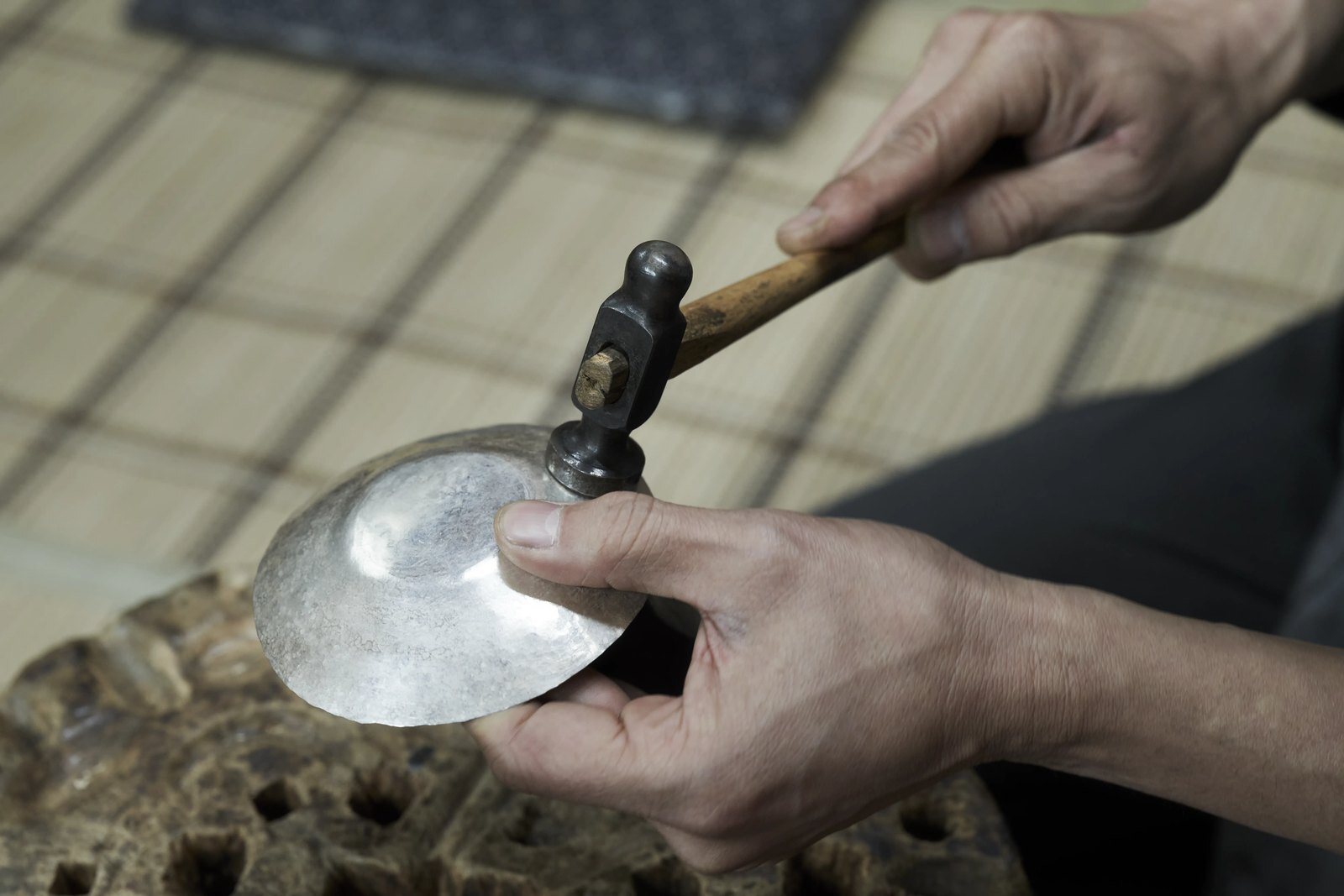 A silversmith's hands holding a small hammer over a shallow hand-raised silver bowl resting on a pitted stake