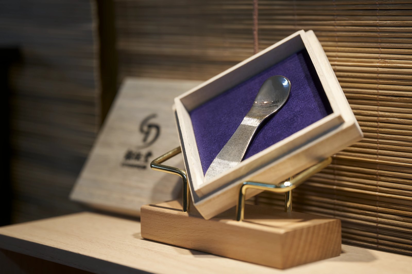 A finished hand-forged silver spoon presented in a purple-lined wooden box, the workshop's mark on the board behind it
