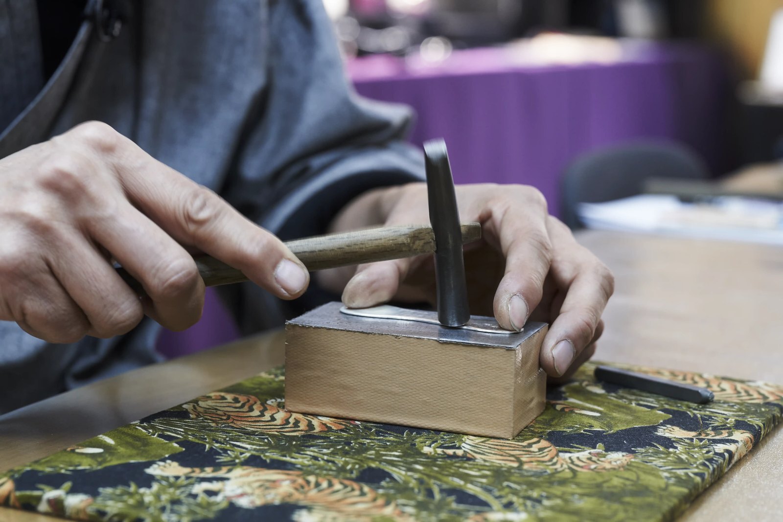 A silversmith chasing a small silver piece with a hand-held chisel and hammer, the work steadied on a wooden block