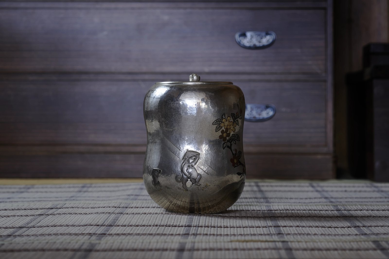 A small hand-raised silver tea caddy with an engraved design, its surface darkened with age, standing on a woven mat