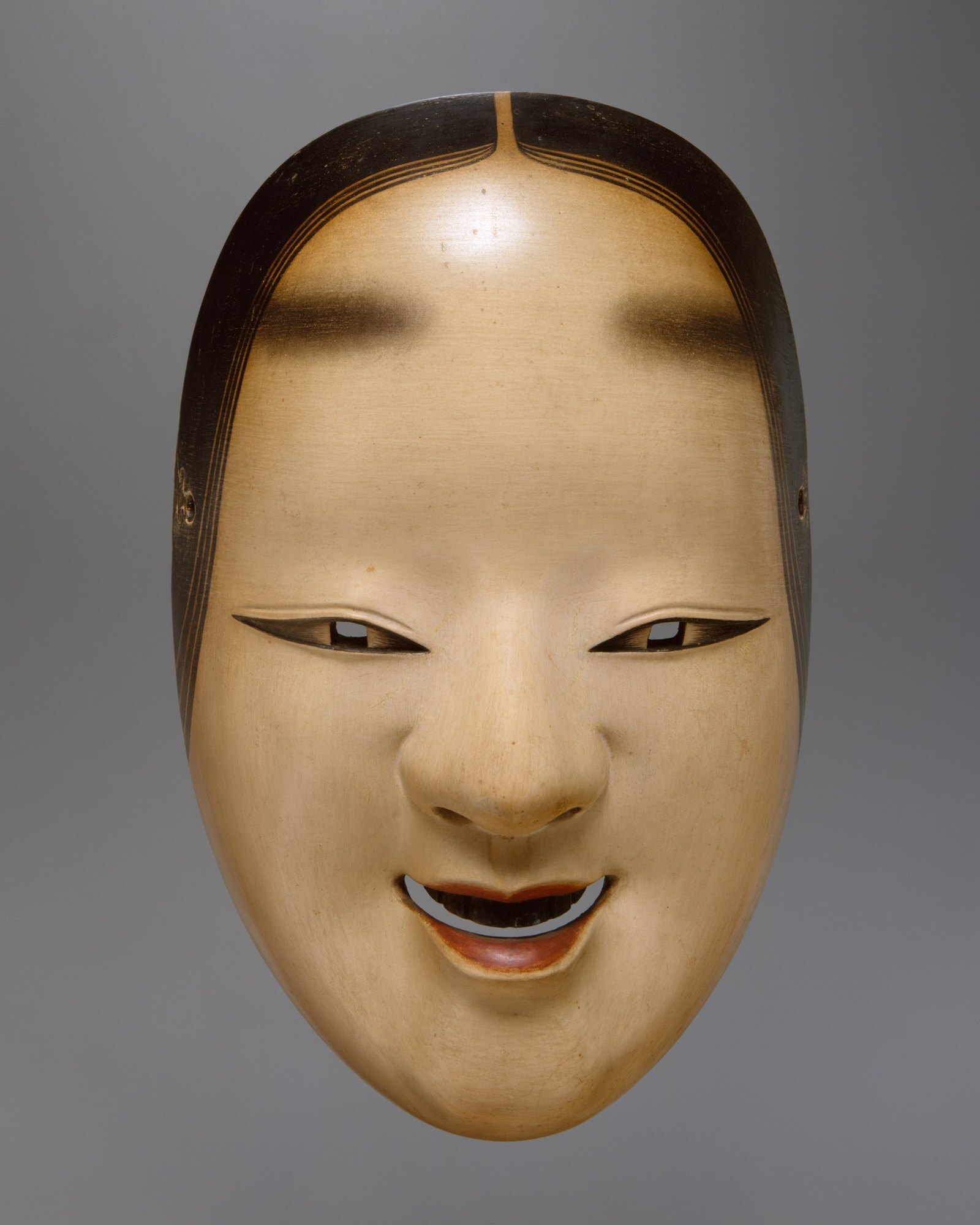 A ko-omote noh mask seen straight on, the carved face lit so that its expression stays undecided