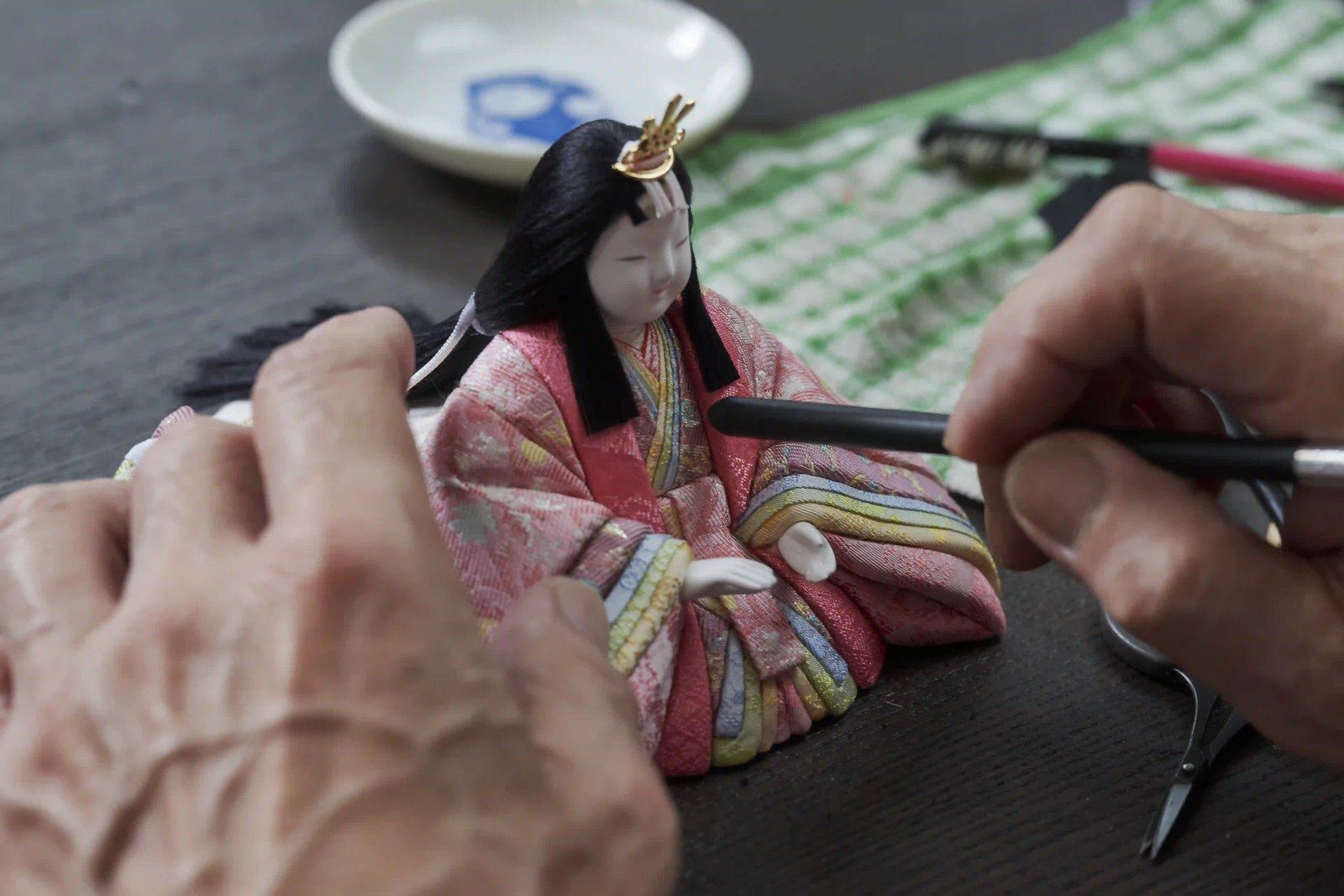 A fine brush painting the features onto a finished doll's face, the dressed body resting on the bench