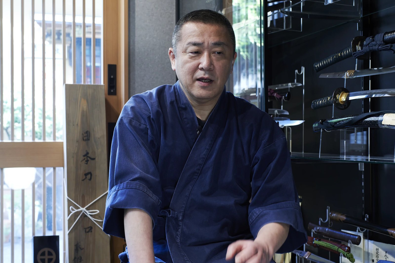 The polisher talking in his shop, a rack of mounted swords behind him