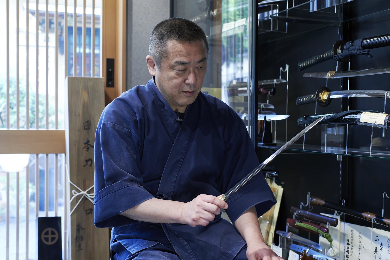 A sword polisher seated in his shop, holding a bare blade up and turning it to catch the light