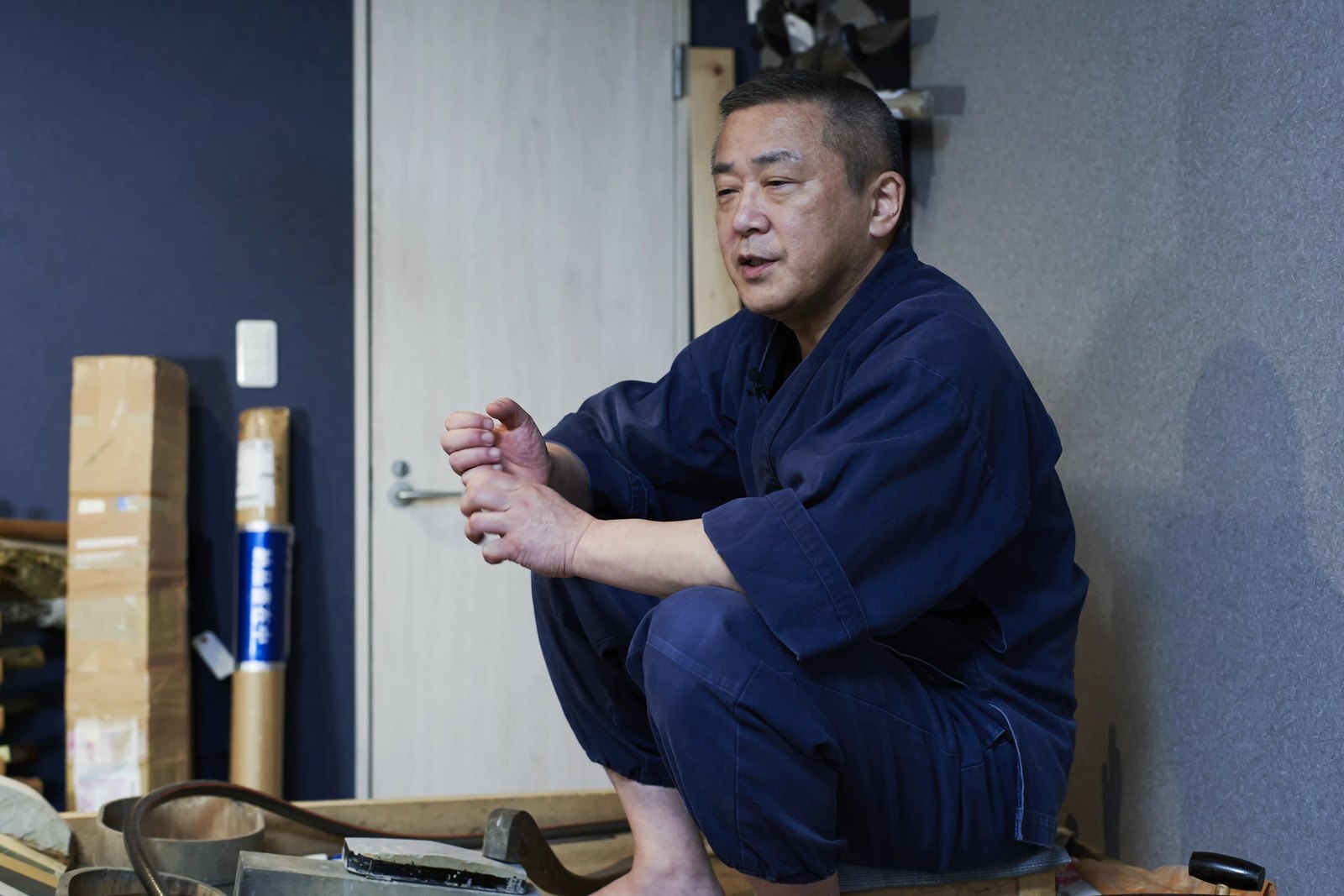 A craftsman in indigo work clothes sitting in his workshop, mid-sentence, one hand raised in explanation, with stone and tools on the boards in front of him