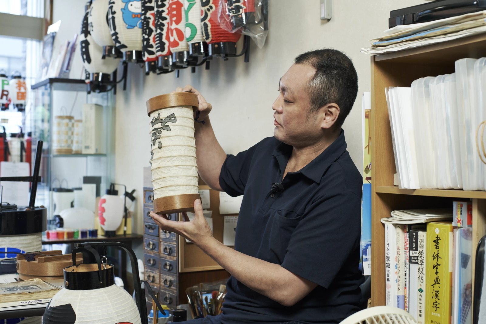 A lantern maker holding a folded paper lantern up in his workshop, finished lanterns hanging on the wall behind him