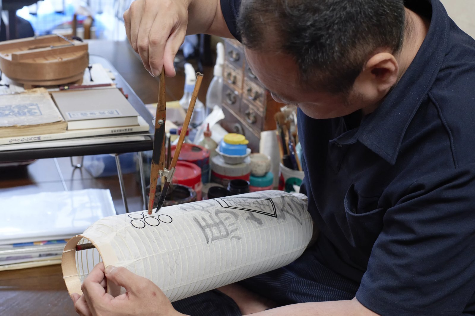A lantern mounted on a wooden stand while the maker works on it, pots of paint set out beside him